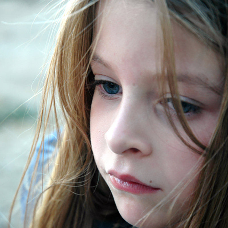 Photo. A young girl looks off camera. She has brown hair blowing in the wind and has a sad / thoughtful expression. She appears to be outside.