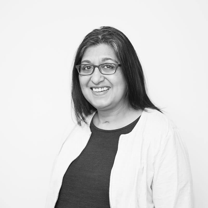 Greyscale headshot of a woman with dark straight hair. She is smiling at the camera, wearing glasses, and a light cardigan over dark top