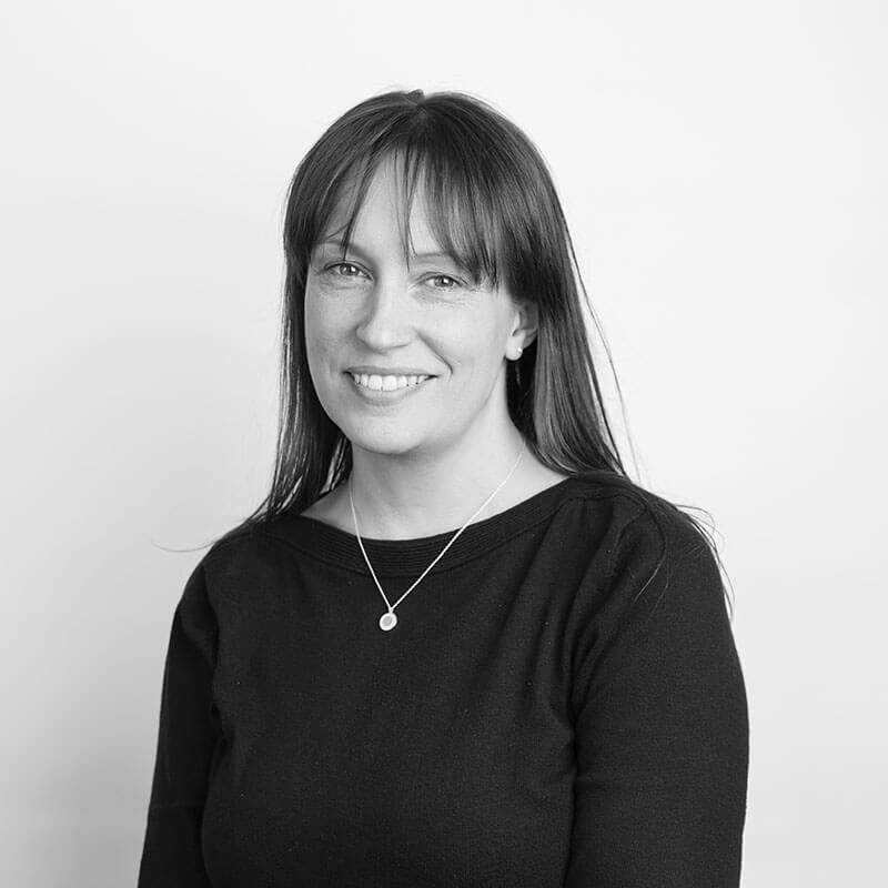 Greyscale headshot of woman with dark straight hair and a fringe. She is smiling at the camera and wearing a dark long-sleeved top.