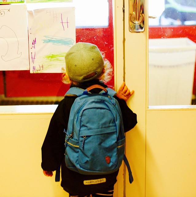 Photo. A child of preschool age with a black jacket, blue bag and green cap stands looking at a nursery notice board. The picture is taken from behind.
