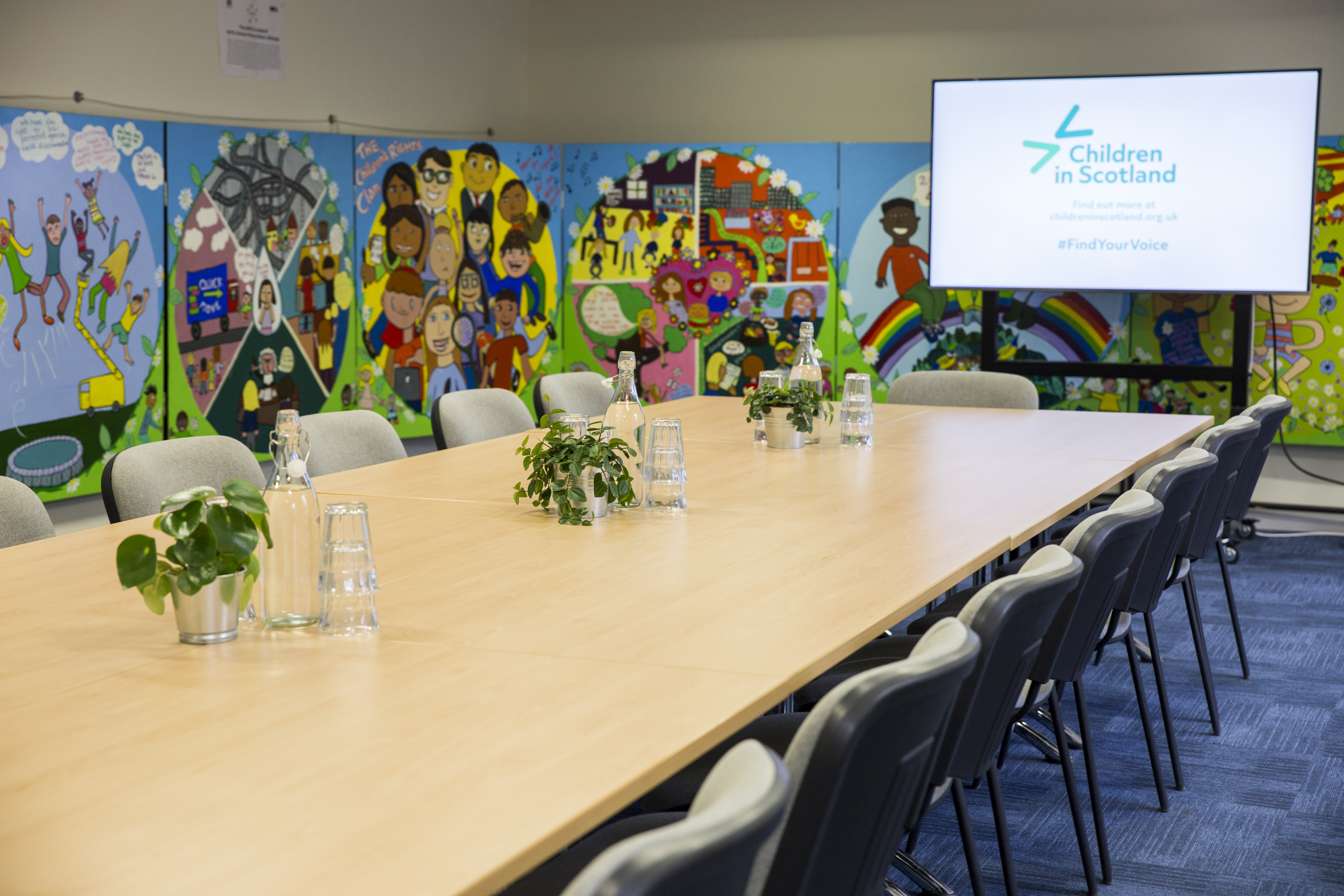 Photo of a large meeting room with a table with chairs on either side of it. At the end is a large screen that says 'Children in Scotland' on it. There are children's drawings on the walls