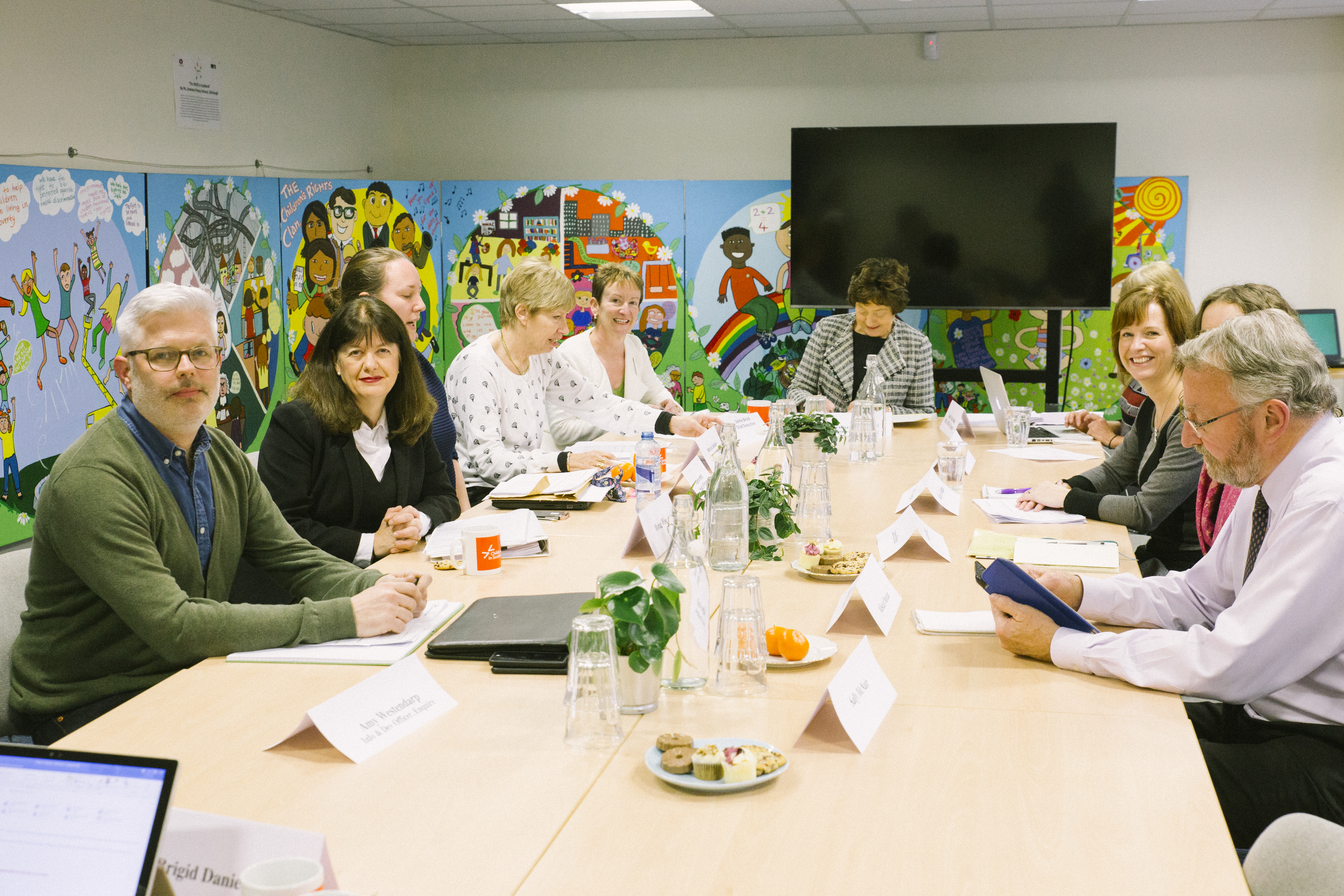 Photo of people sitting around a meeting table. Some of them are looking at the camera while others talk. There is a large screen at the back of the room and the walls are covered in children's drawings