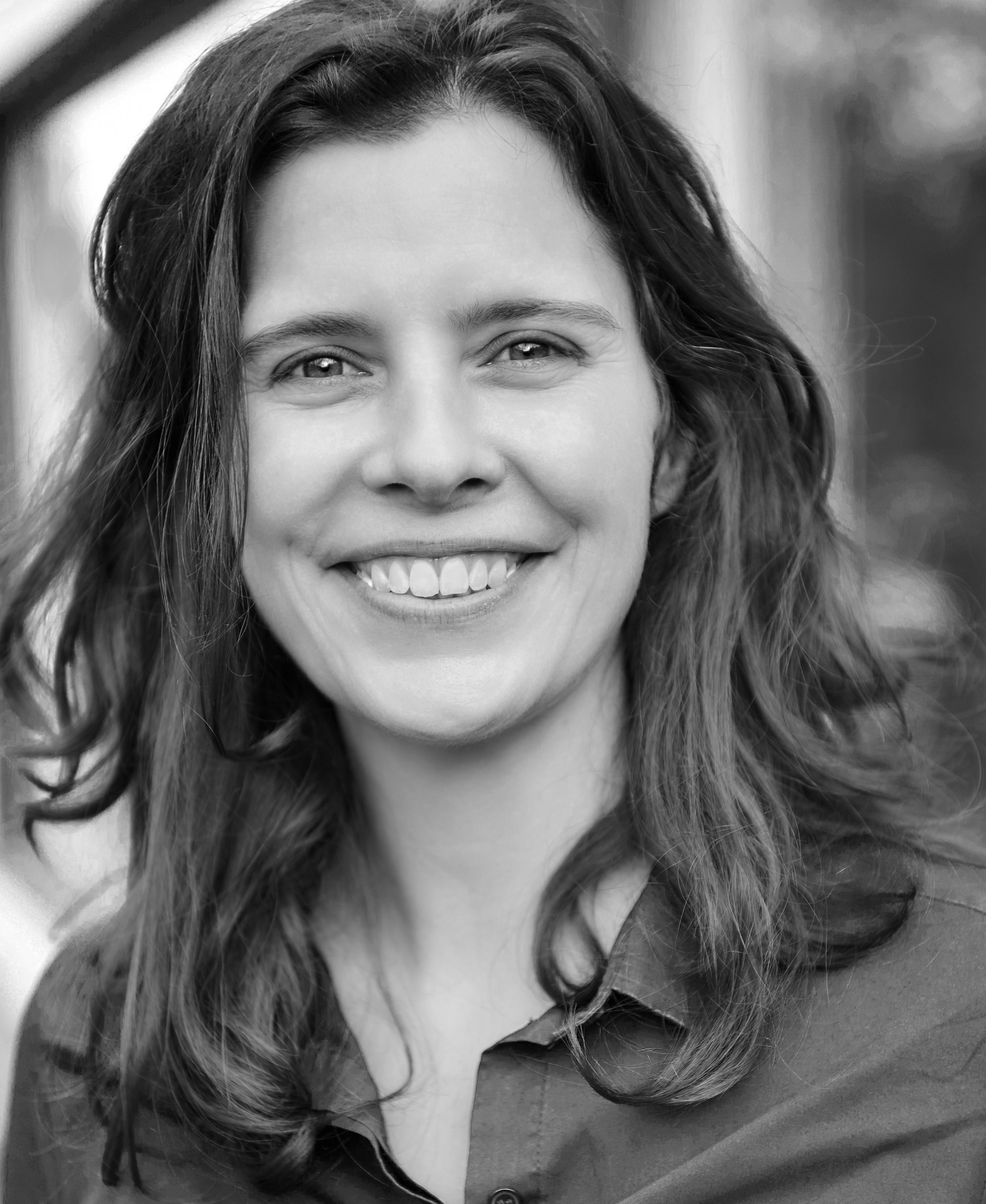 Greyscale headshot of a woman with dark long wavy hair. She is smiling at the camera and is wearing a shirt.