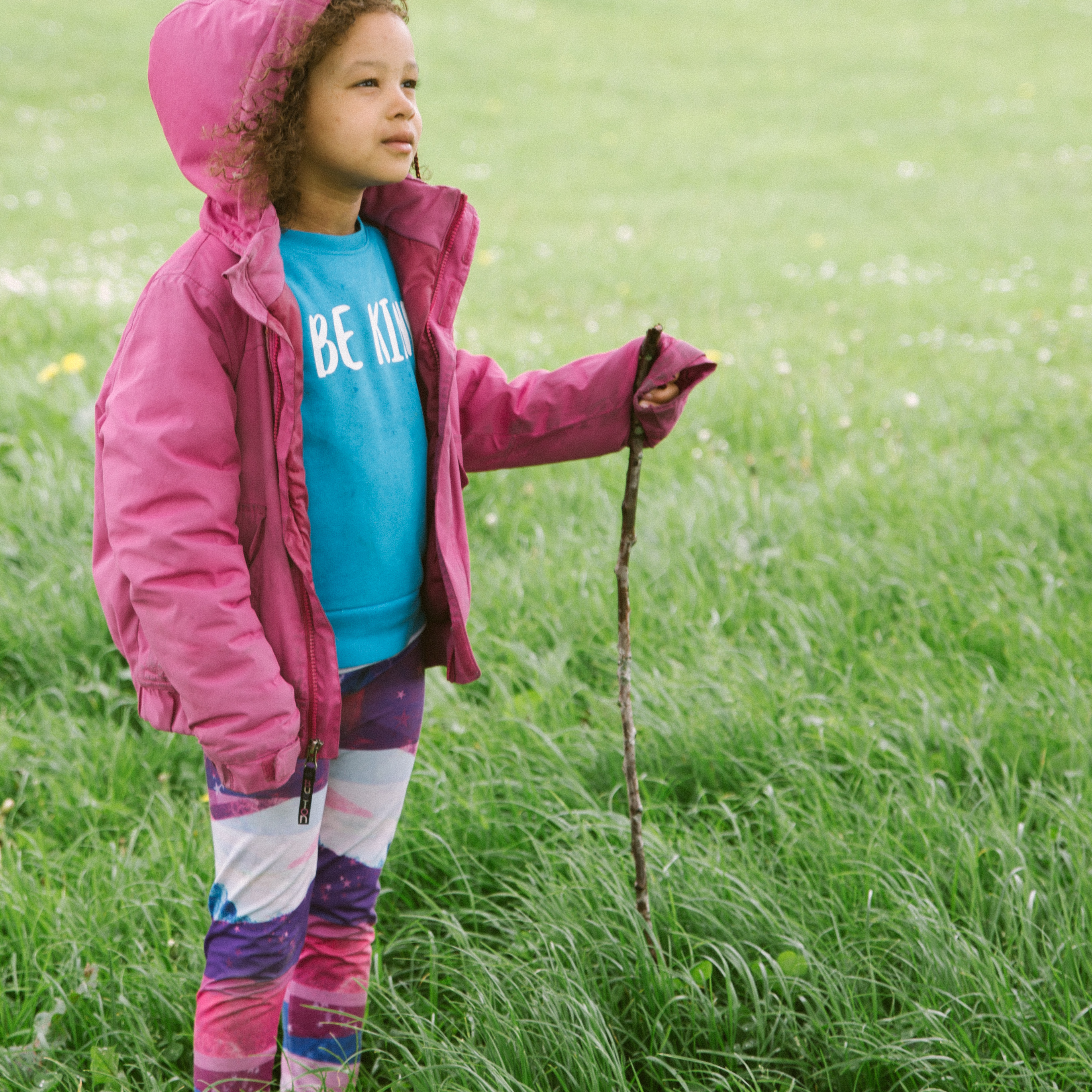 A photo of a young child standing on some grass, she is wearing a pink coat with the hood up and is looking off towards the right.