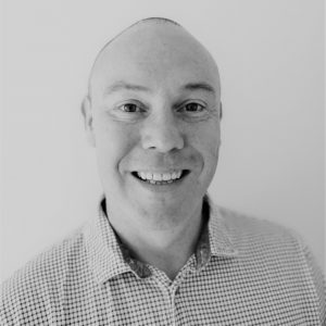 Greyscale headshot of a man smiling at the camera. His hair is short and he is wearing a shirt