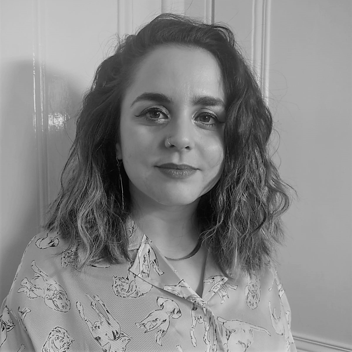 Greyscale headshot of a woman with shoulder-length dark wavy hair. They are looking at the camera and wearing a floral shirt.