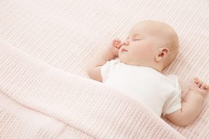 A baby sleeps on its back, arms raised. It is tucked under a pink blanket