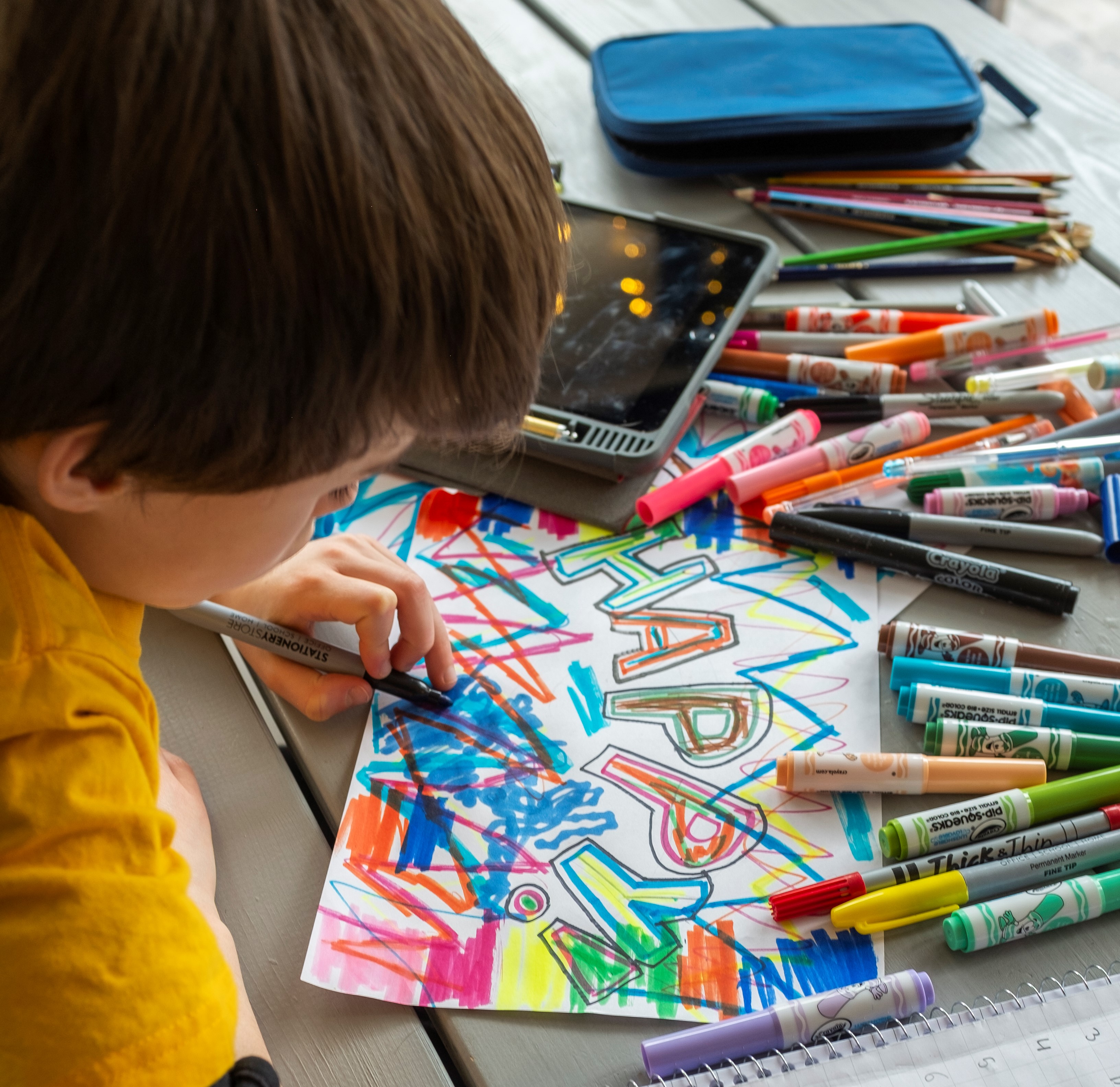 Photo of young person rawing on a piece of paper with a marker. The paper says happy! and there are other coloured markers scattered on the table