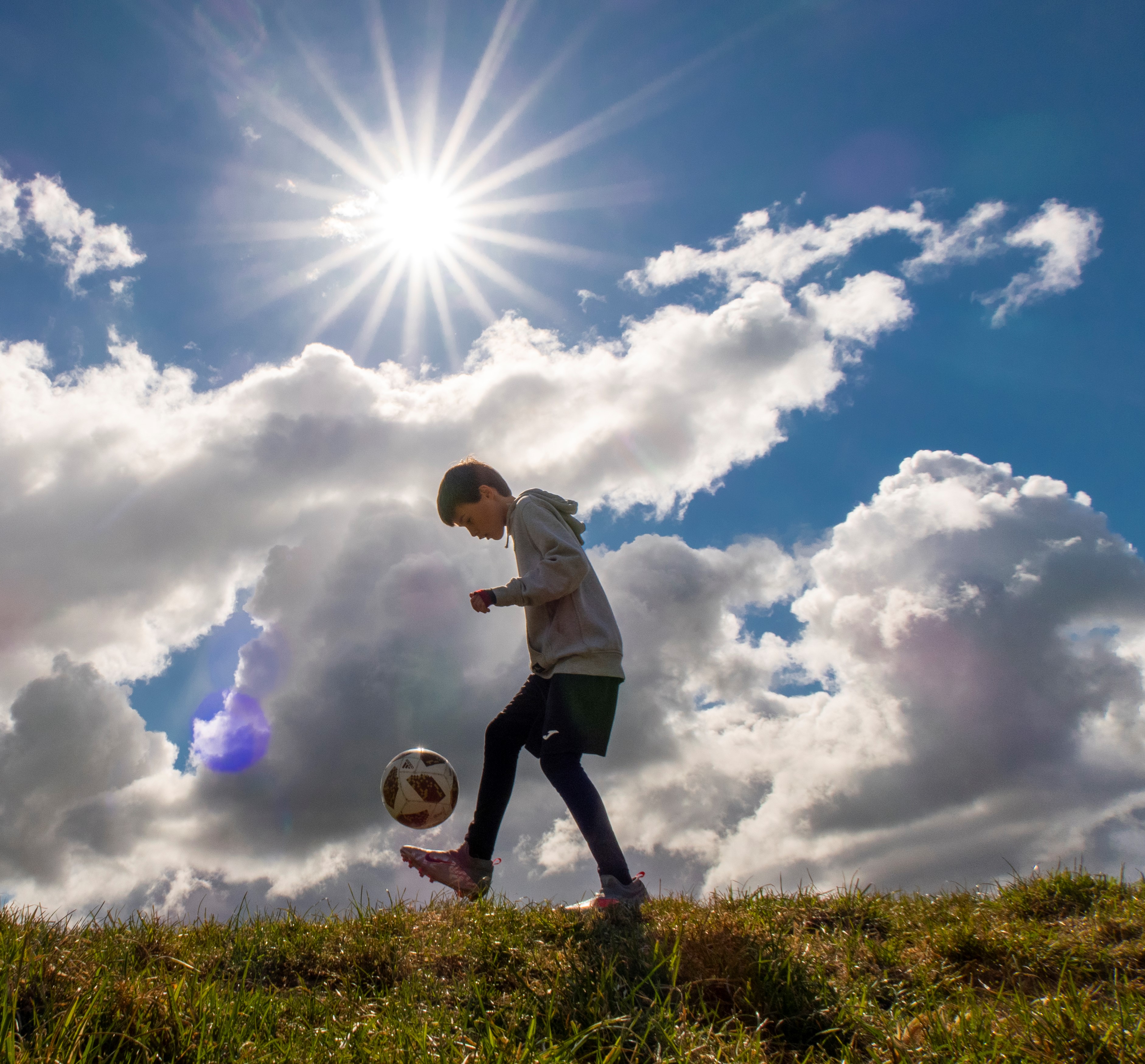 Photo of young person kicking a football on some grass, wearing football clothing. The sky is big and blue, with some clouds and the sun is bright