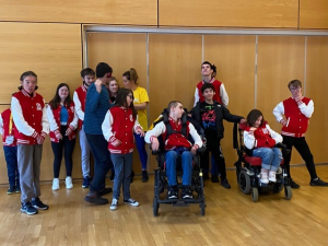 A group of young people wearing jeans and white and red varsity jackets. The floor is wooden and the wall behind them is wood