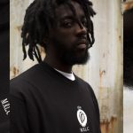 A black man from the chest up, looking at the camera. He has short dreadlocks, a beard, and is wearing a black tshirt that has a white icon and text that reads MRLC