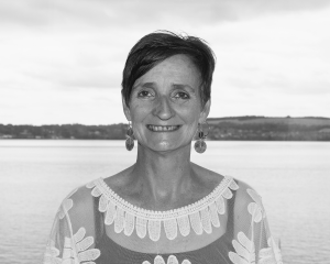 Black and white headshot of a woman with short dark hair. She is smiling at the camera. She is wearing dangly earrings, a white lace top, and is standing in front of the water, with a town visible on the other side.