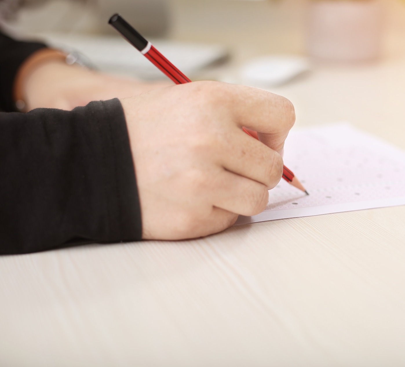 Photo of hands holding a pencil and filling out a paper exam on a light wood table