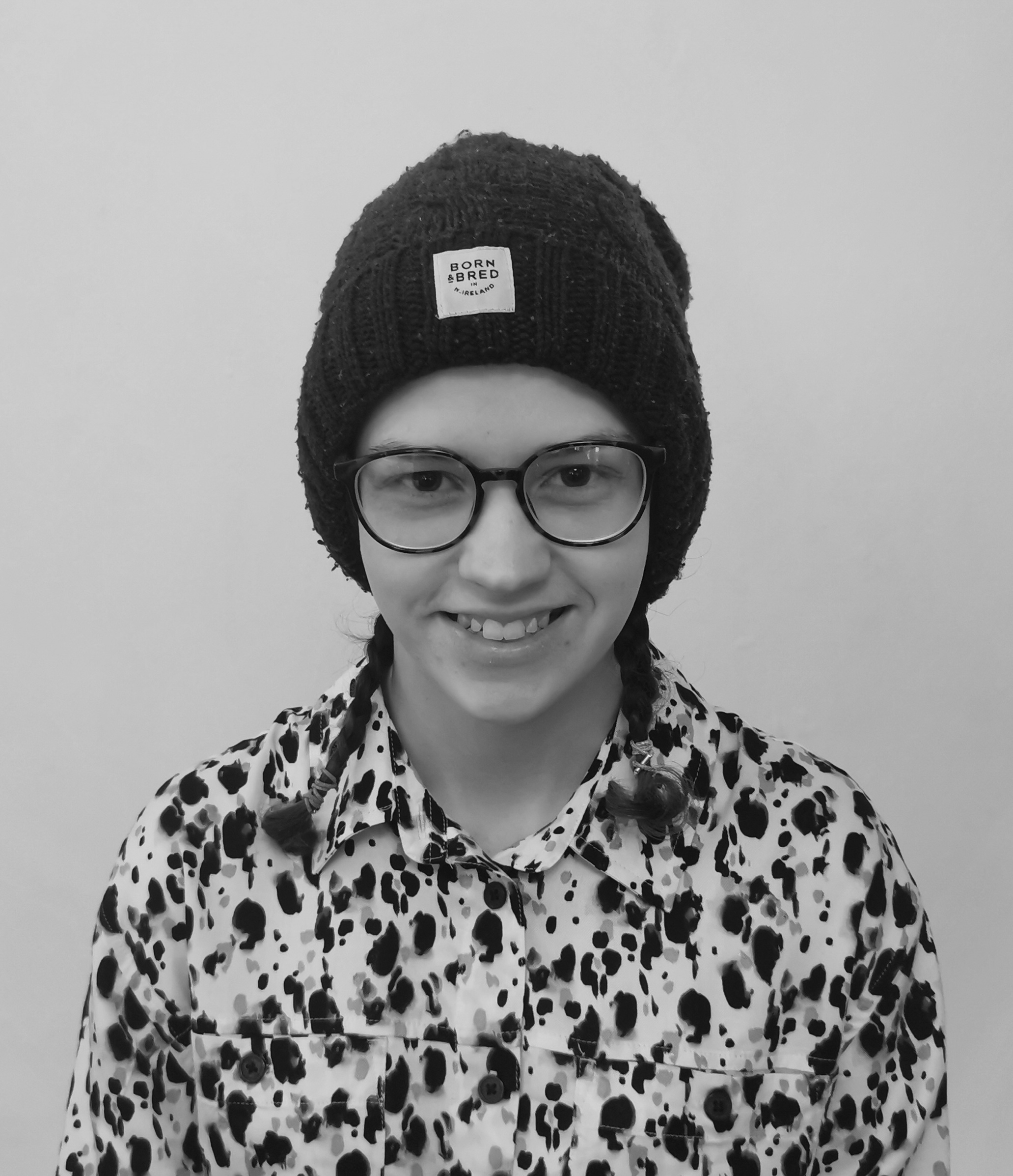 A black and white image of a person from the chest up, wearing a shirt. They have two plaits and are wearing a hat and glasses.