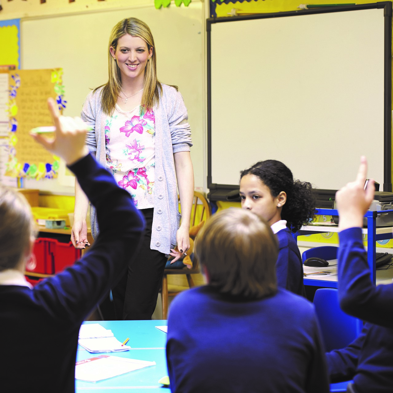 Teacher in class room with school pupils raising their hands.