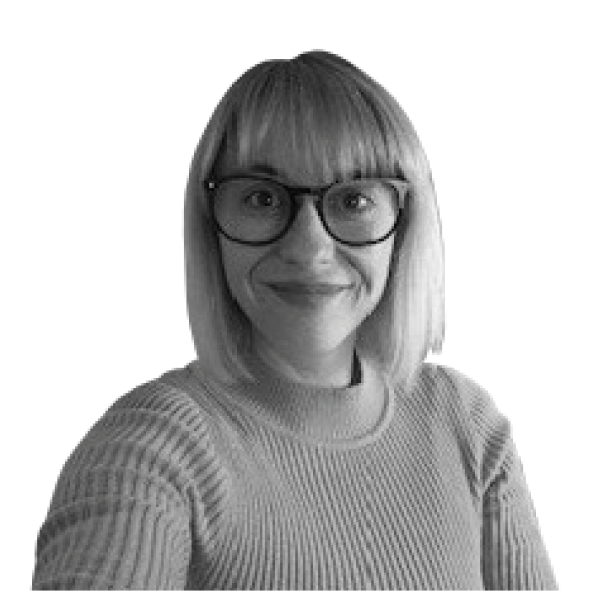 A black and white image of a woman smiling from the shoulders up. She has blond straight hair in a bob with a fringe and is wearing glasses and is looking at the camera.