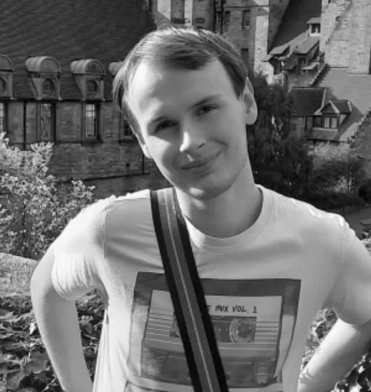 A black and white image of a young man from the upper body upwards. He is wearing a graphic tshirt and has a bag strap across his body. He has light brown hair and is smiling at the camera.