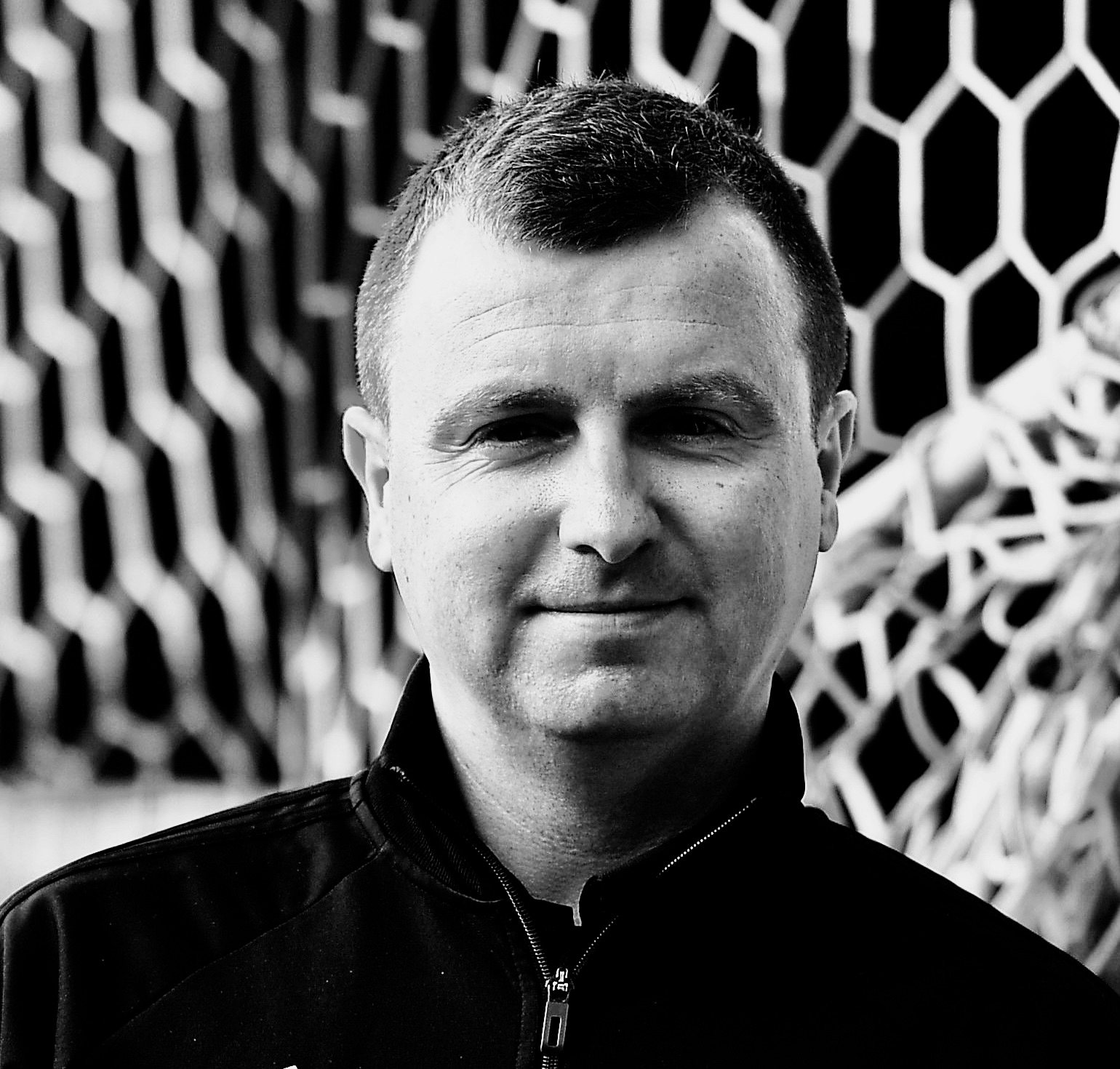 A black and white image of a man looking directly at the camera from the shoulders up. He has short dark hair and is wearing a dark zip-up jumper. In the background there is hexagonal netting.