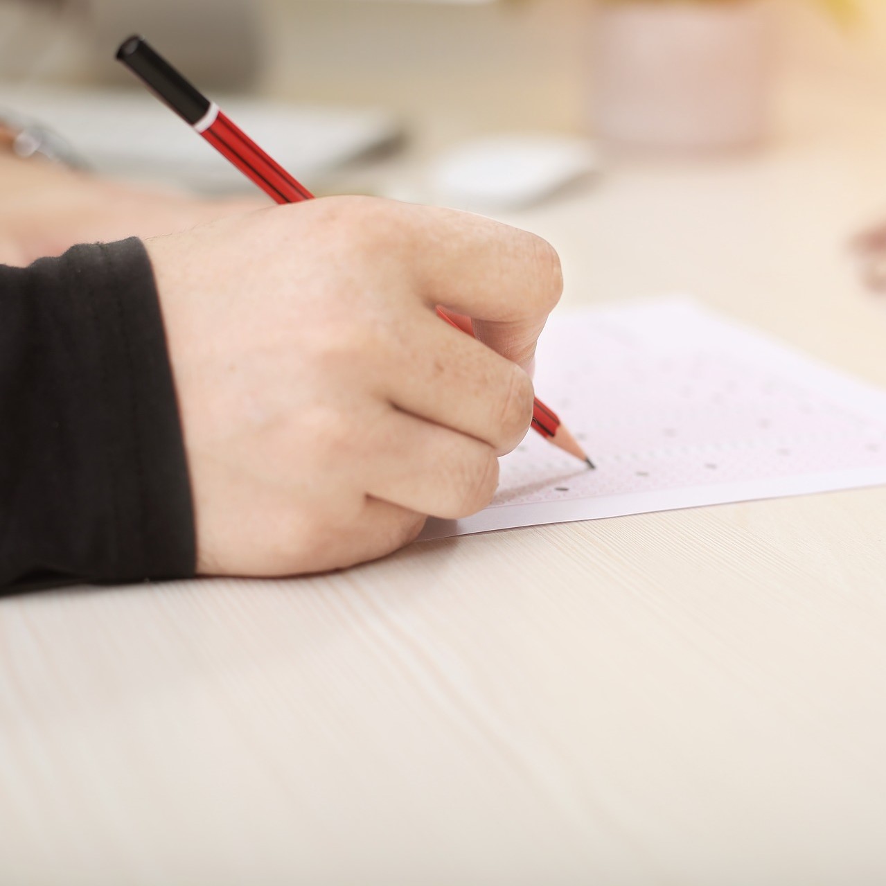 A hand holding a pencil filling in an exam questionnaire on a light desk