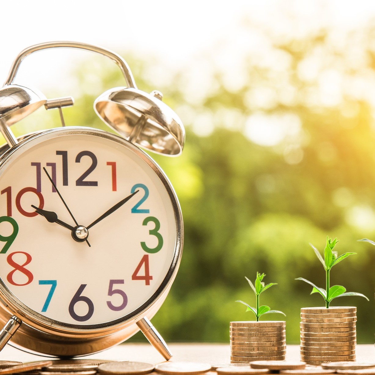 An alarm clock with large colourful numbers next to coins. Some of the coins are in two piles, with plants growing from the top.