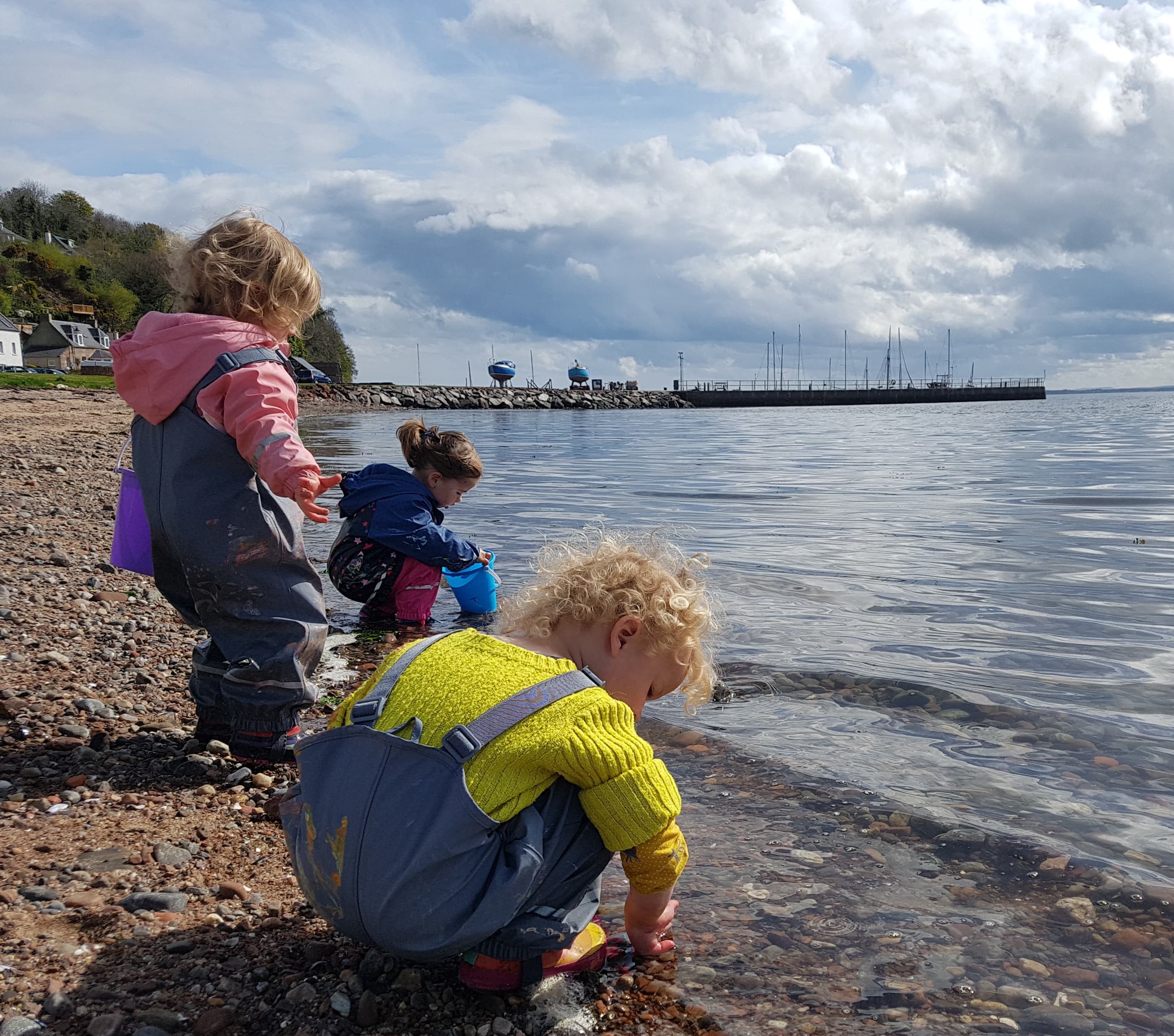 Young children of pre-school age explore a stretch of pebble beach by the water. A young boy in yellow is crouched at the water edge.