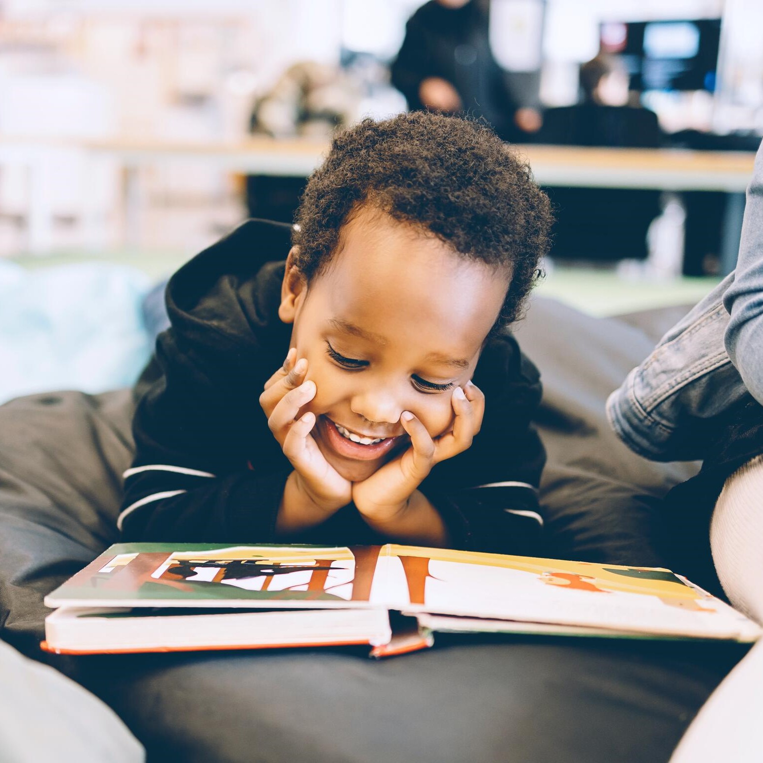 A photo of a young boy reading a picture book. He is lying on his front with his hands over his cheeks, and is smiling.