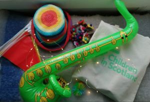 A photo of an inflatable green saxophone, a Children in Scotland canvas bag, some bunting, and a rainbow knitted hat