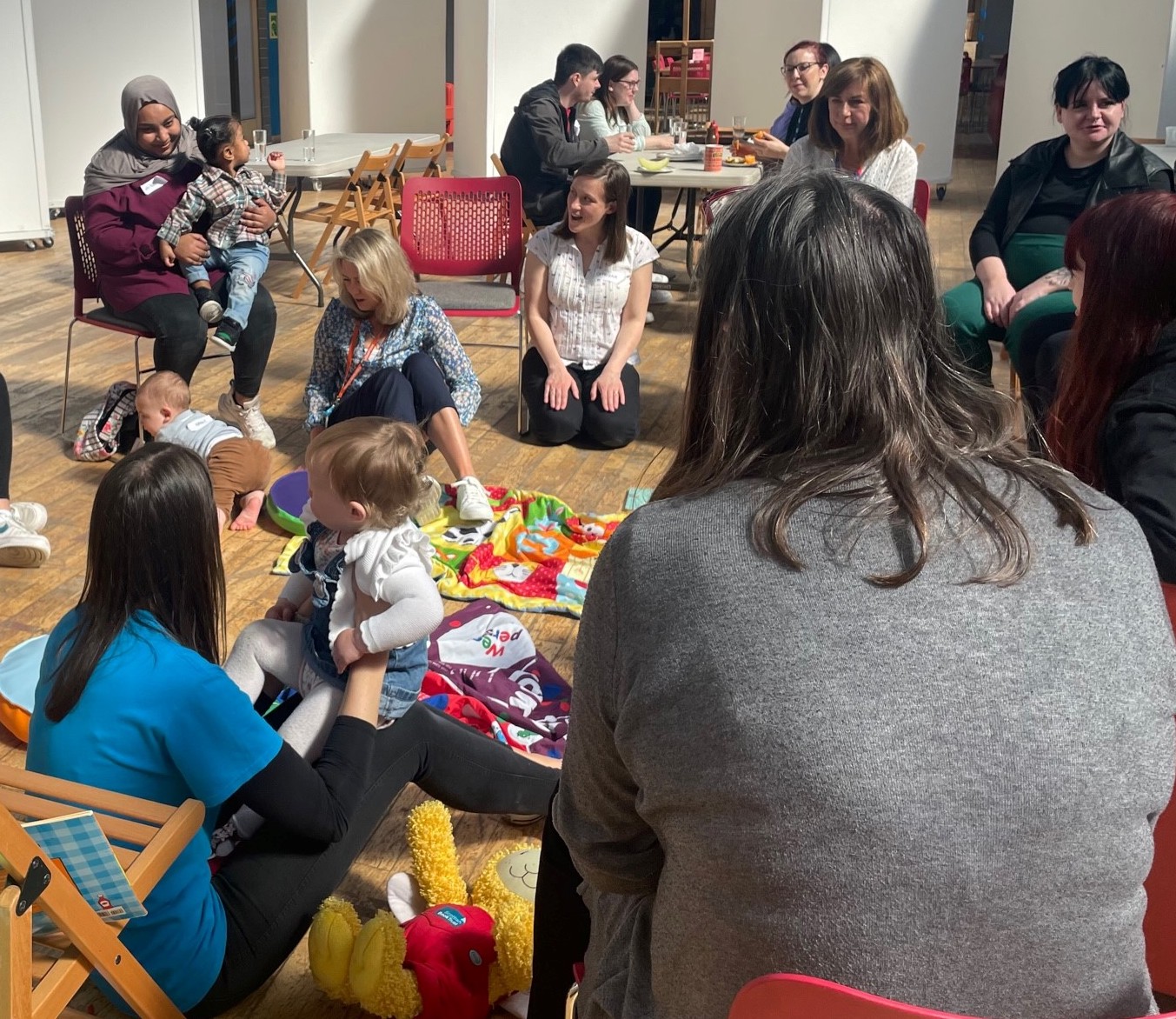 A photo of children and adults in a playgroup. They are sitting down on the floor and on chairs, talking to one another. There are toys and play things on the ground.
