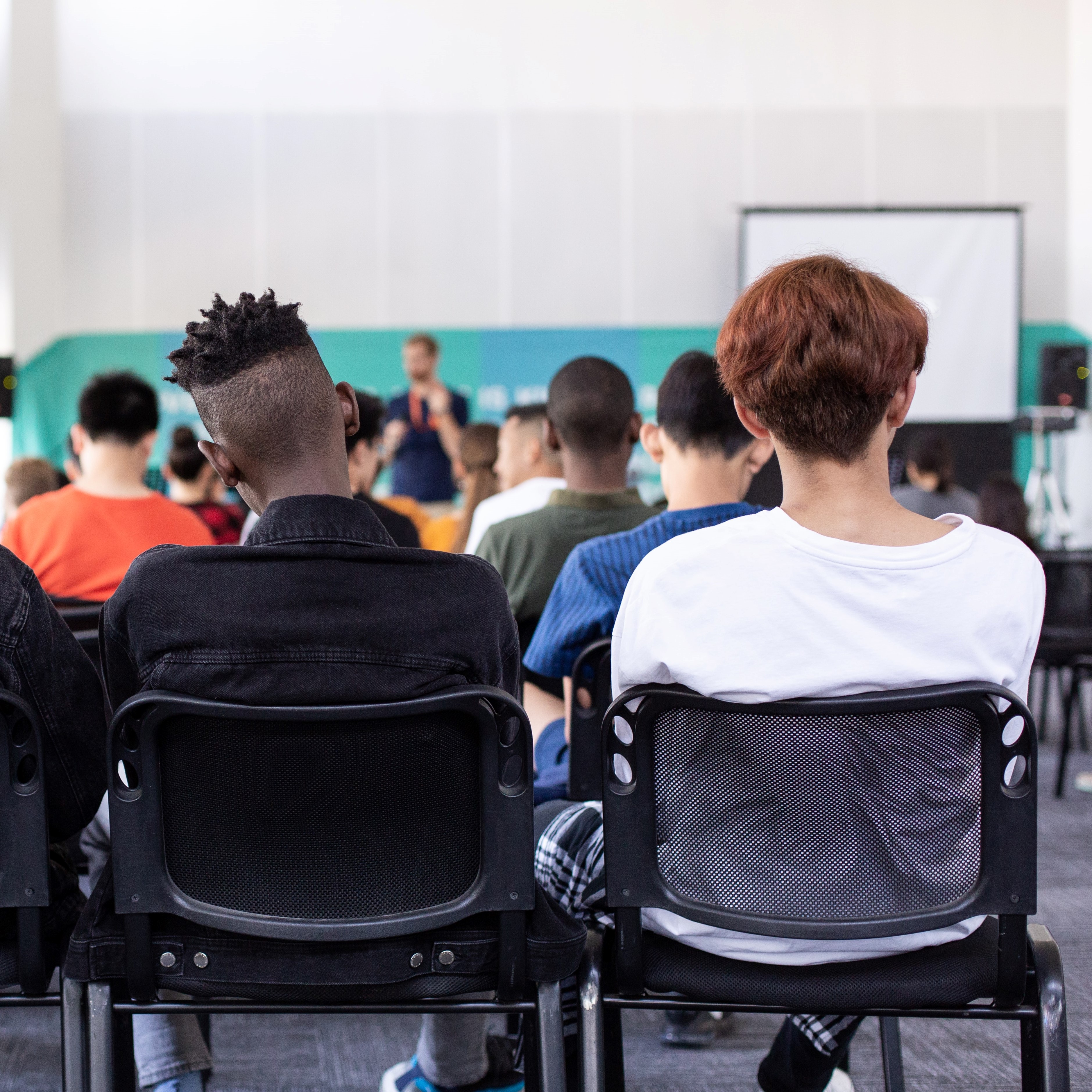 A photo of young people sitting in chairs facing away from the camera and towards a speaker at a whiteboard.