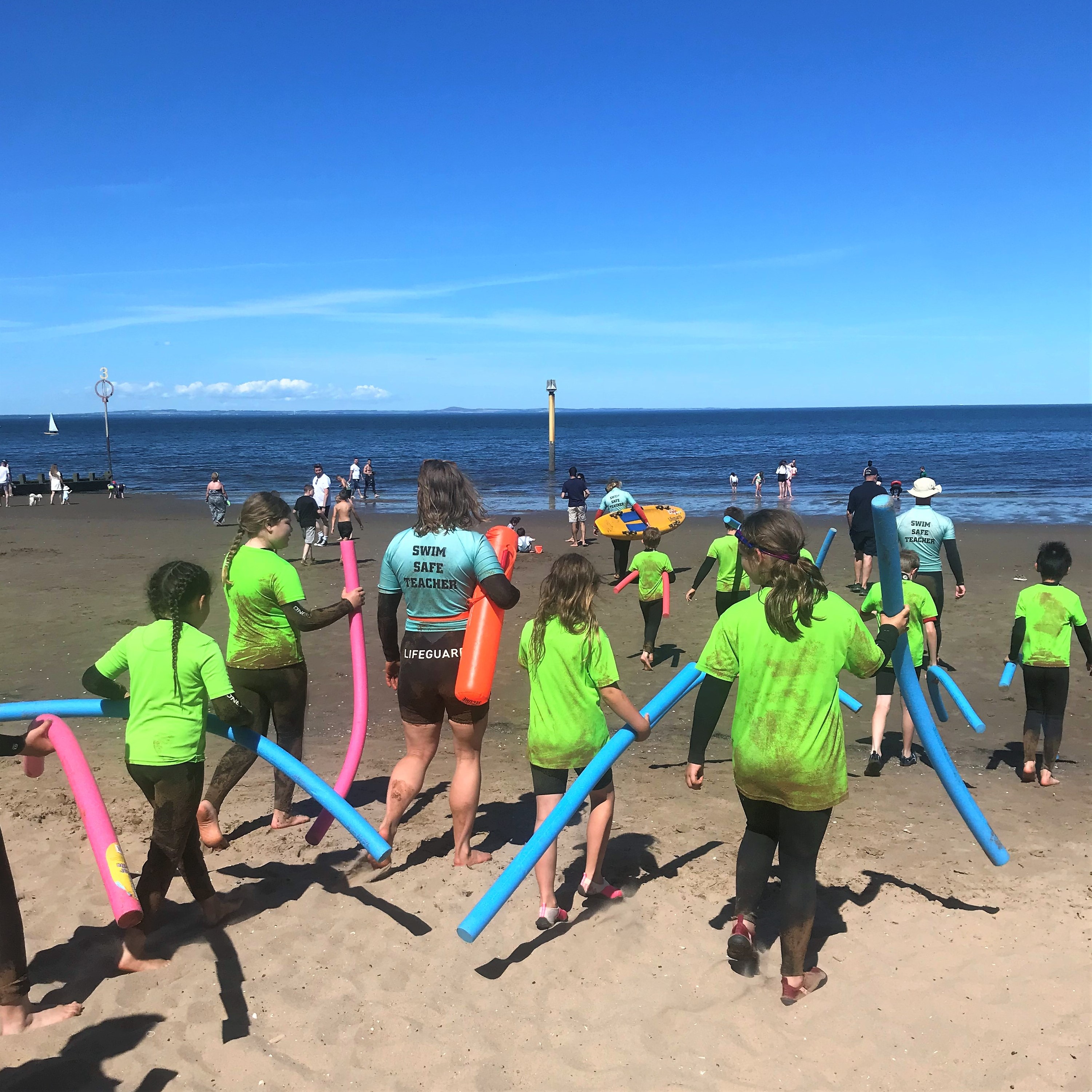 Photo of children on a beach holding long water floats. An adult is wearing a t-shirt that says 'swim safe teacher' on the back.