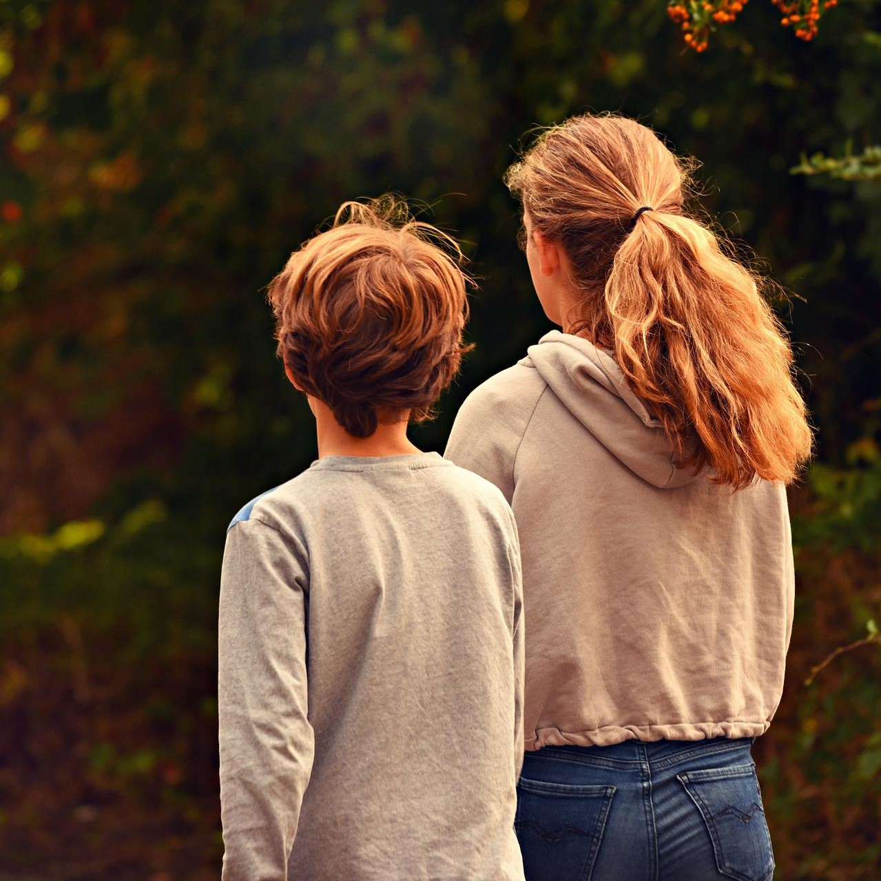Photo of two children walking away from the camera. The one on the left is shorter, with short hair, and the one on the right is taller and has long hair in a ponytail.