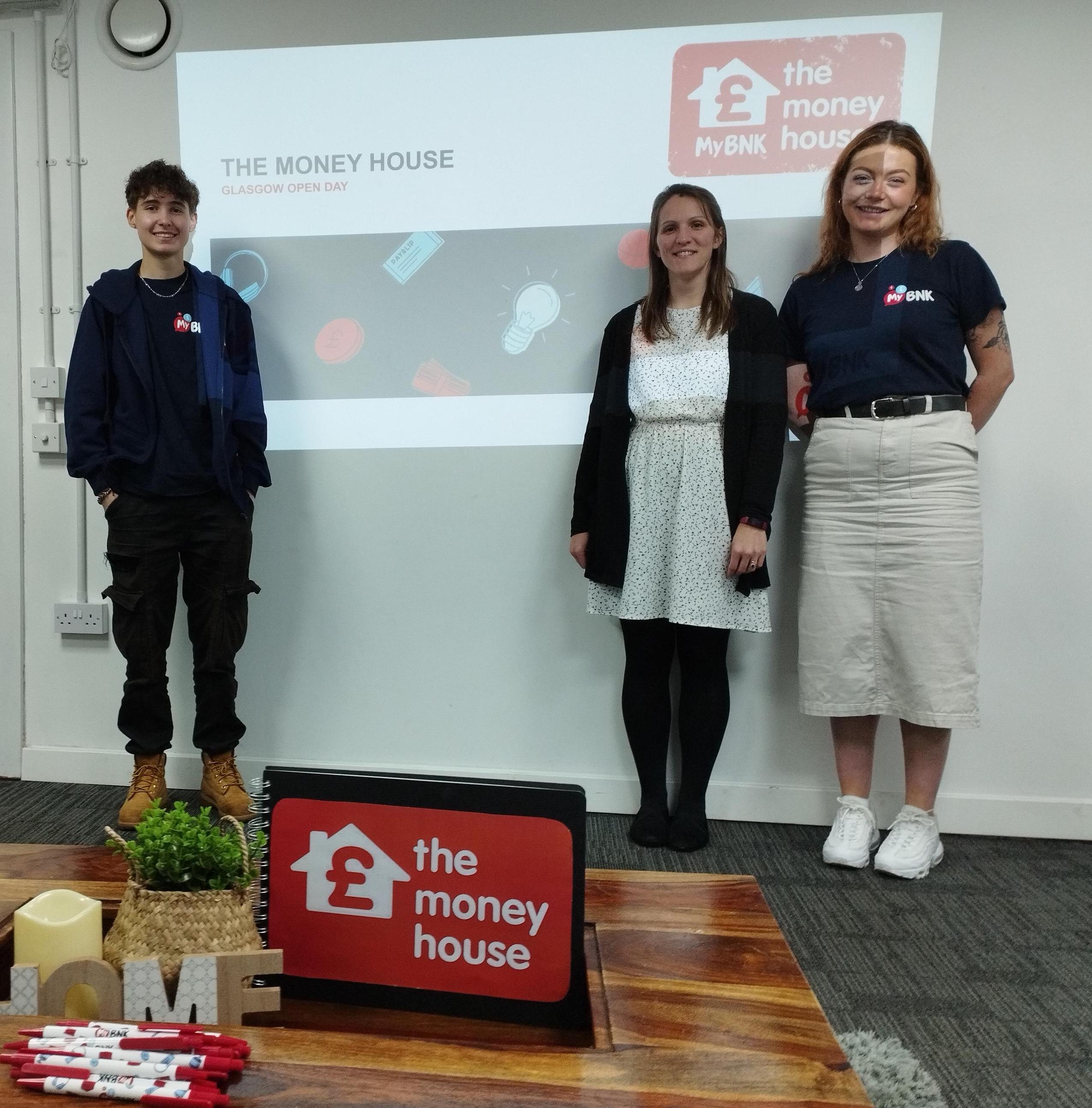 Three young people stand in front ion a projector screen which shows 'The Money House logo and the words The Money House Glasgow Open Day. At the foreground is a wooden table with a plant, wooden lettering spelling out 'Home', and Money House branded material such as pens.
