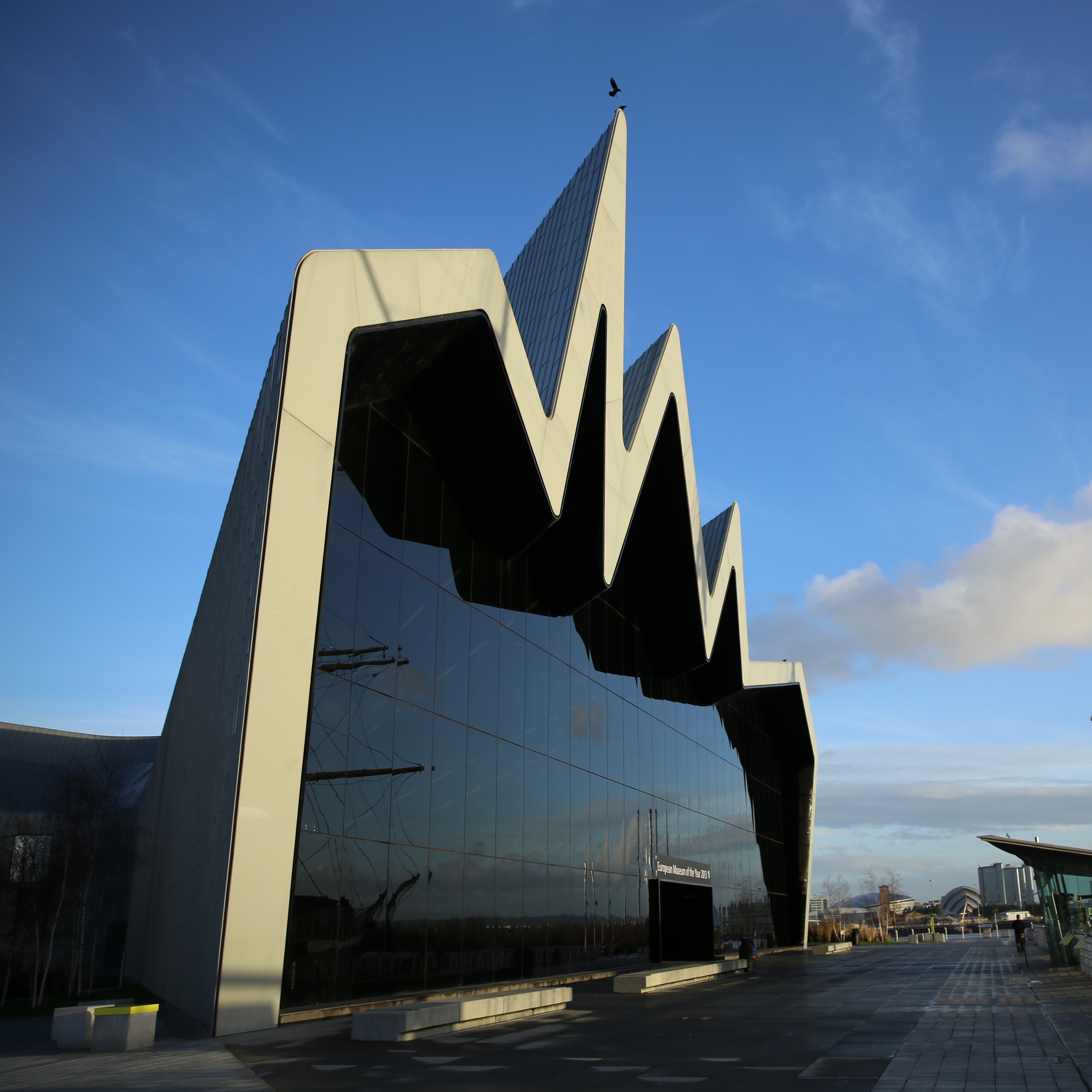 Photo of the Riverside Museum with a blue sky in the background. The building's visible wall is entirely glass, with a jagged metal framework outline.