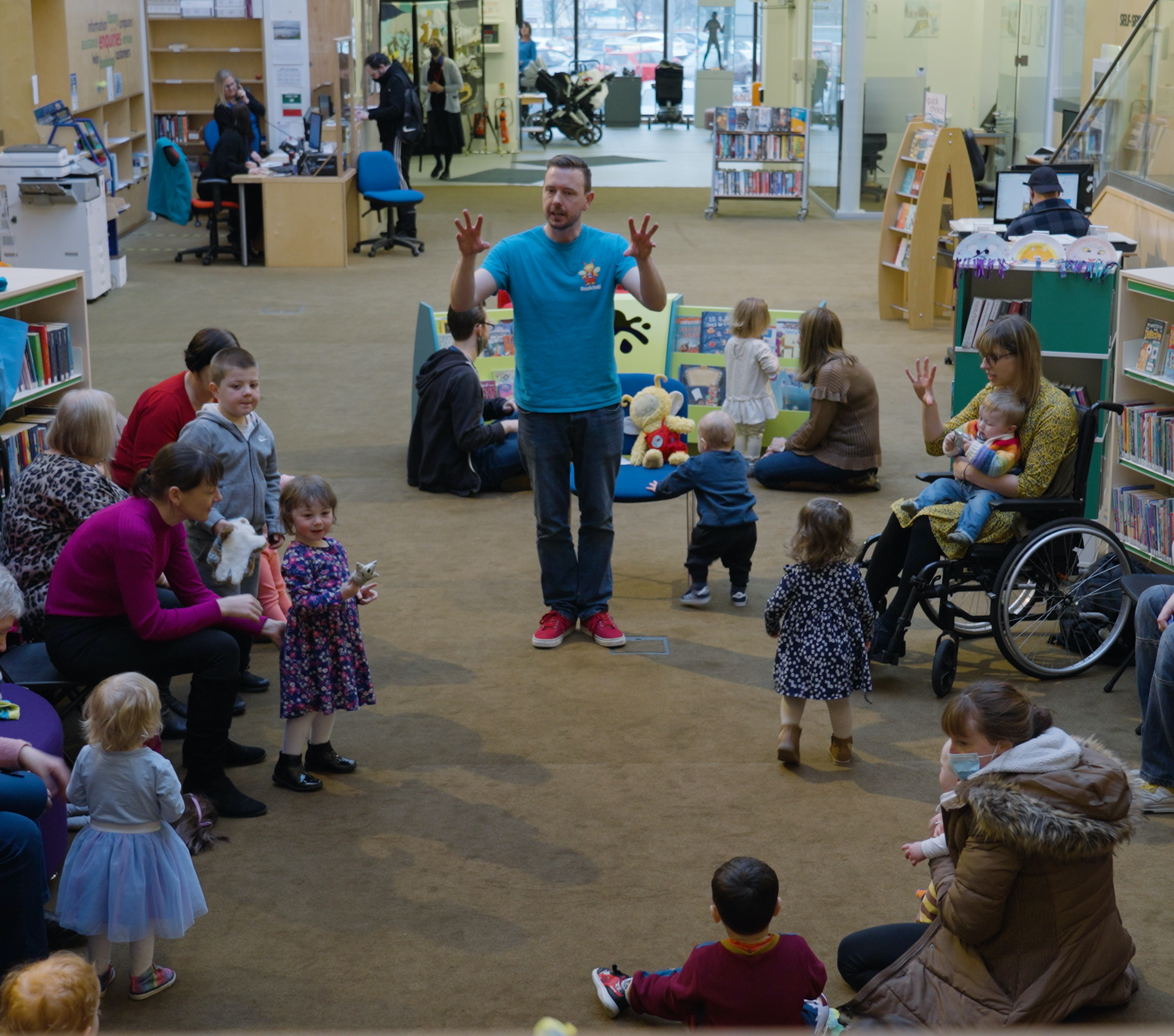 Photo of an adult running a Bookbug session in a library. A man in blue tshirt and jeans stands amongst a group of pre-school children, toddlers and their parents and carers. He is leading them in song.