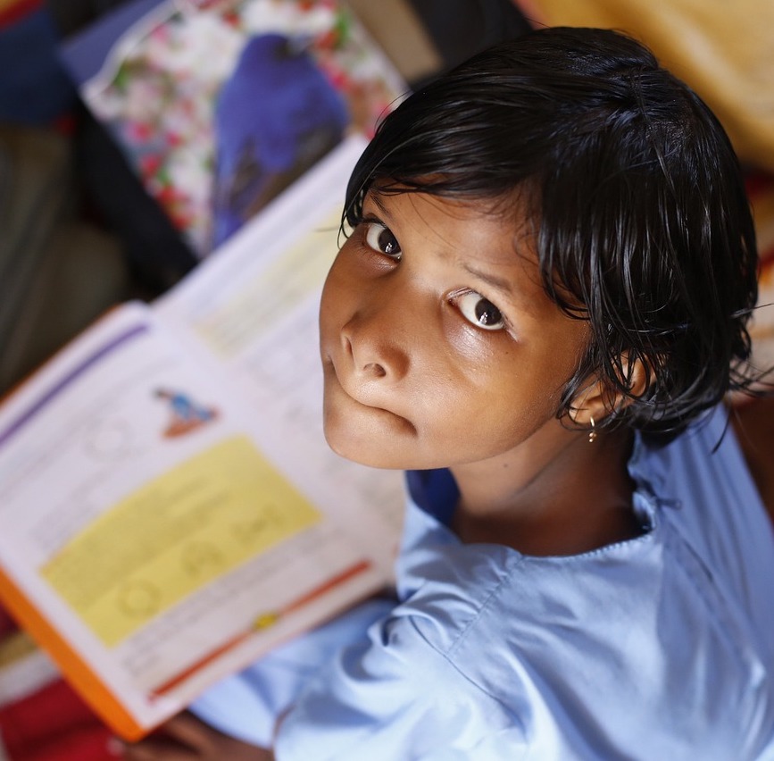 Photo. A young school-aged girl sits with an open textbook looking over her shoulder and up to the camera. She has brown skin, black hair and is wearing a light blue top.