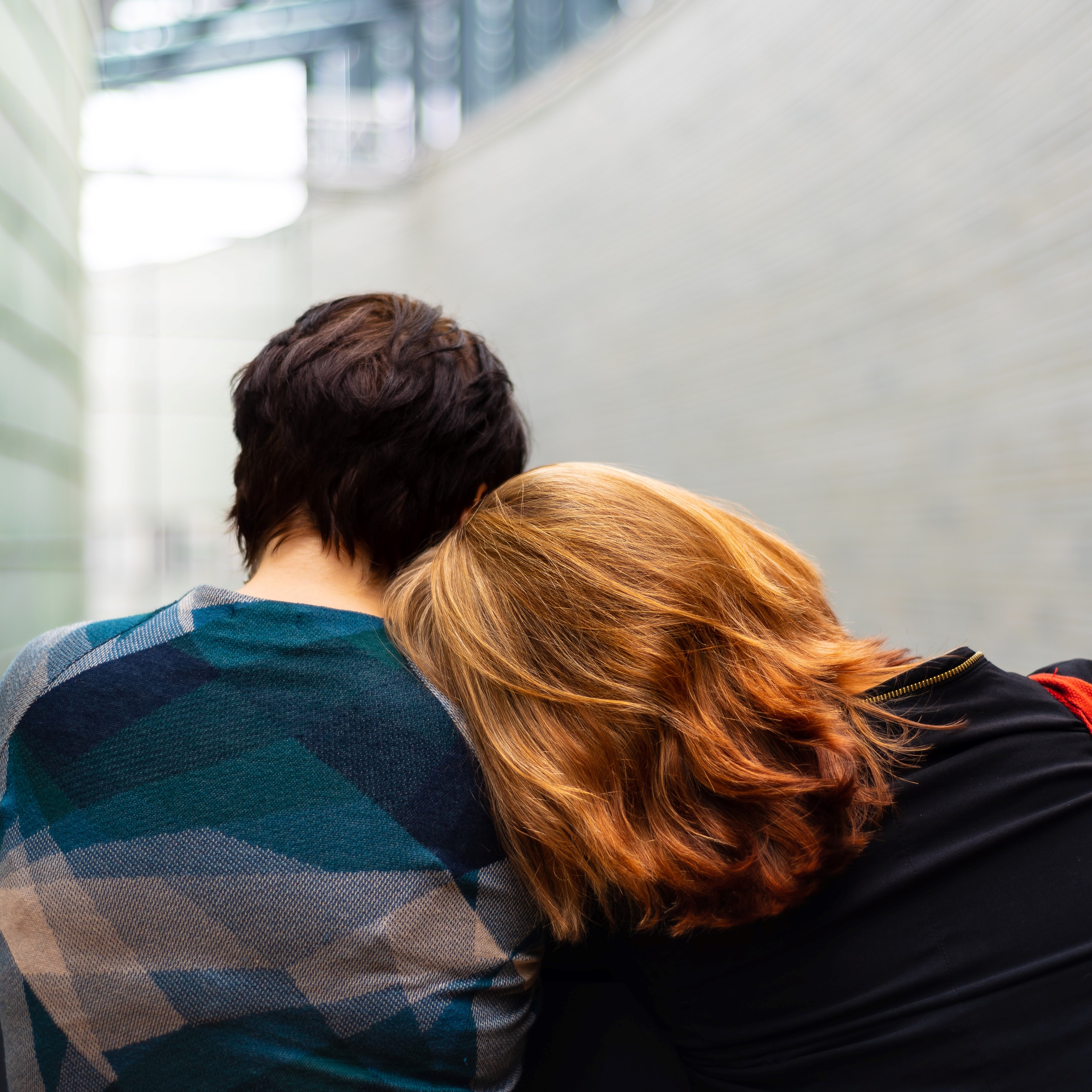 Two people with their backs to the camera. One has short dark hair, the other is leaning against them and has long orange hair.