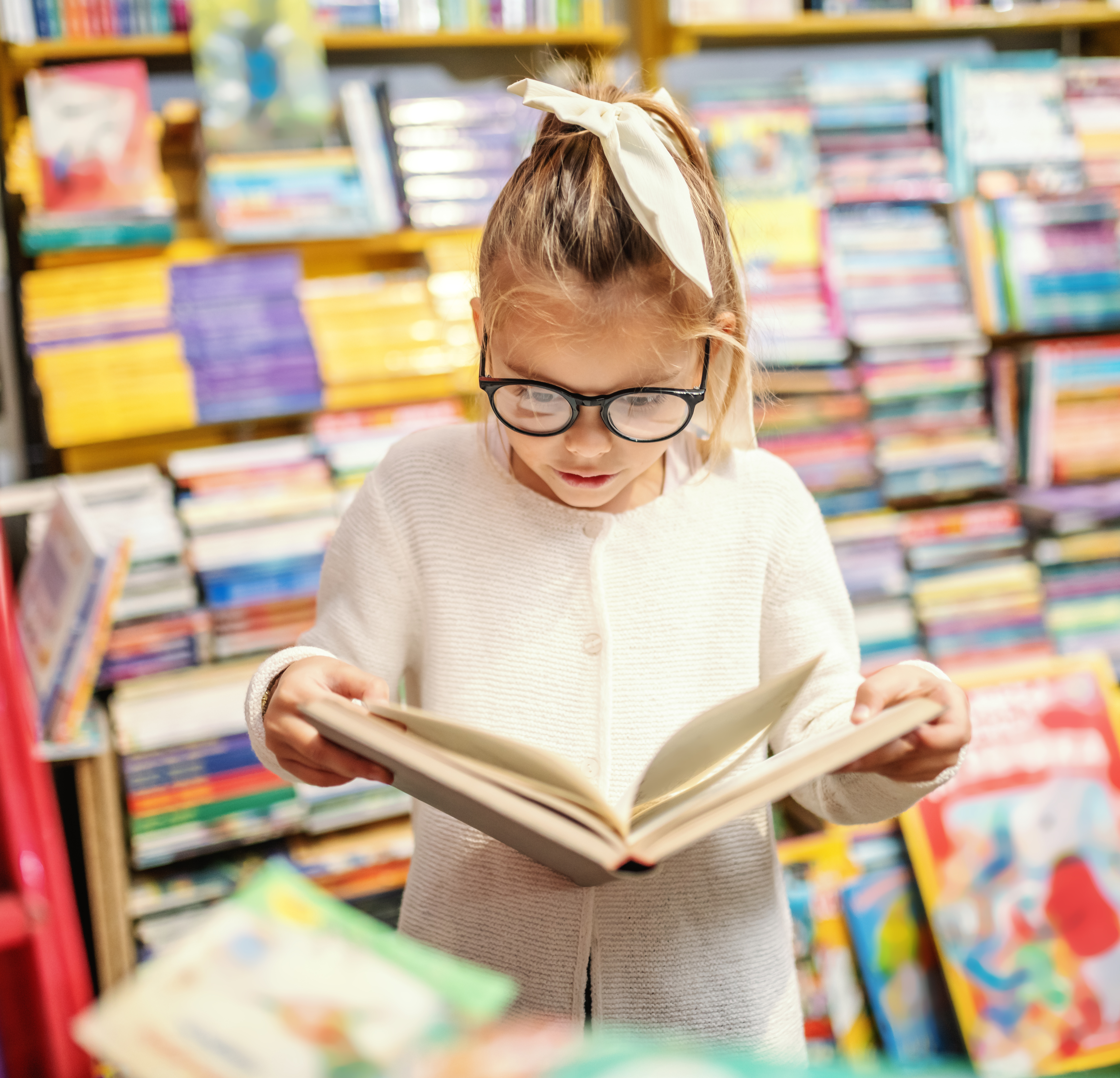 A little girl with glasses stands with an open book. Behind her are shelves full of books and there are some out of focus in the foreground. 