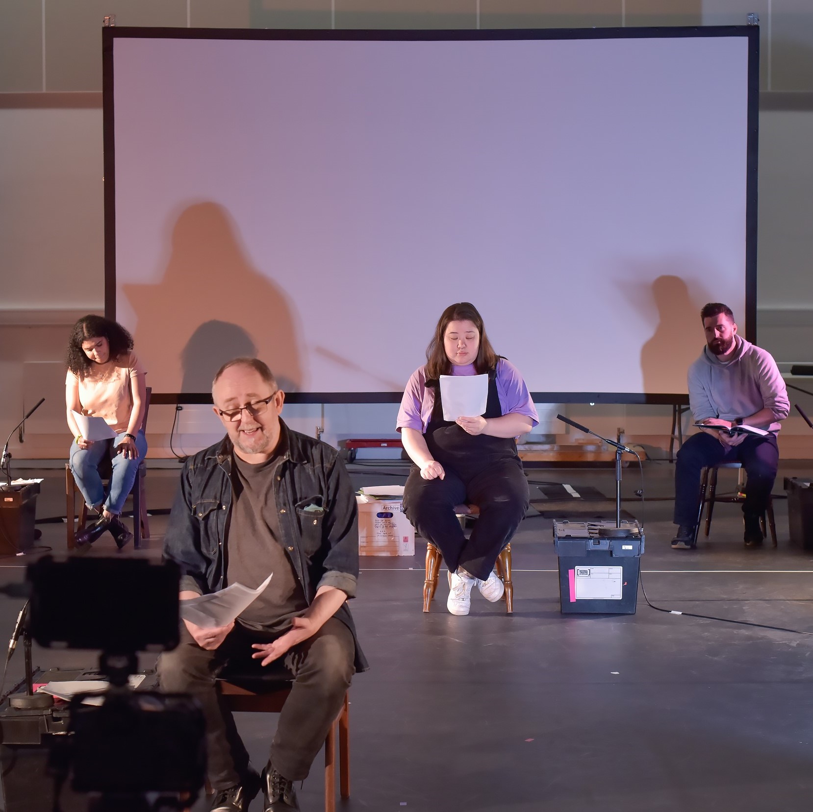 Photo of four people sitting on chairs reading from paper. There is a white projector screen at the back, and a filming camera in the foreground.