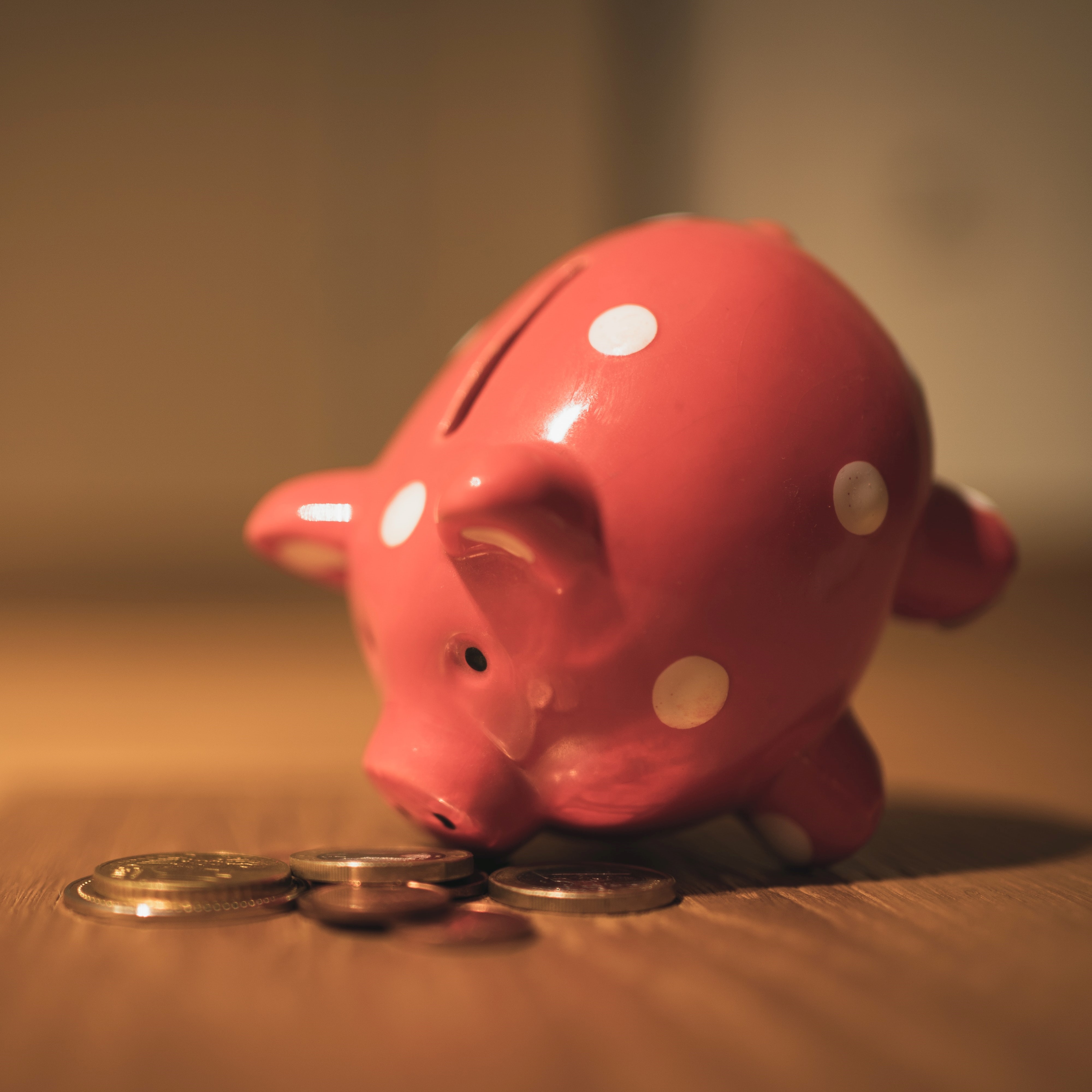 A pink piggy bank leaning on its nose, with coins surrounding it on the table