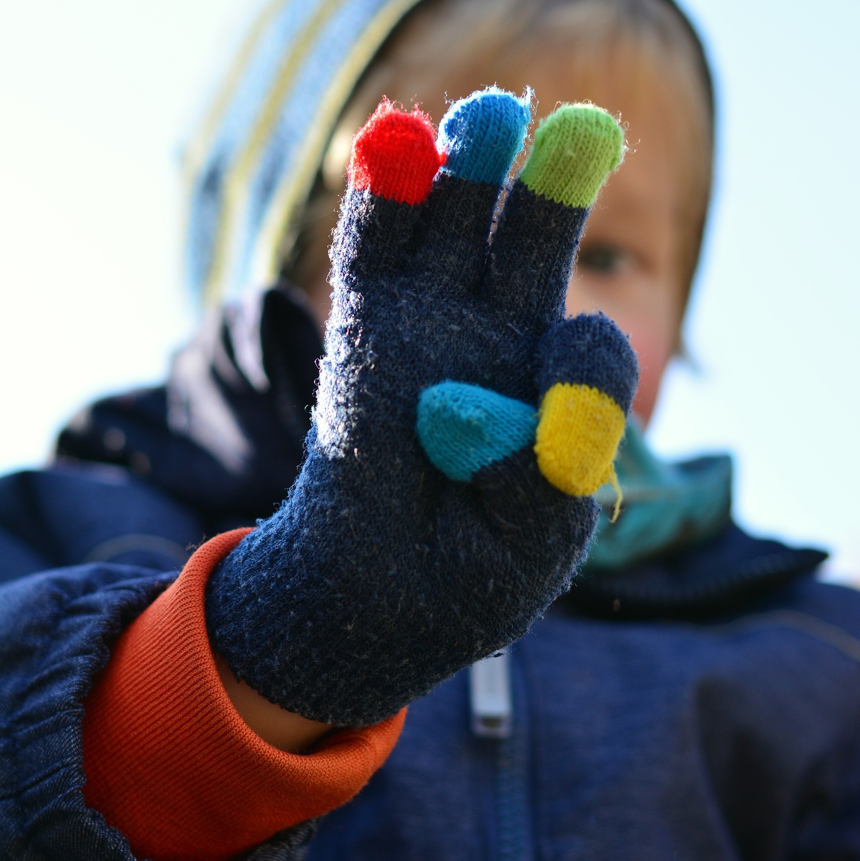 A young child with their hand extended towards the camera, with three fingers up. They are wearing woollen gloves and a hooded coat.