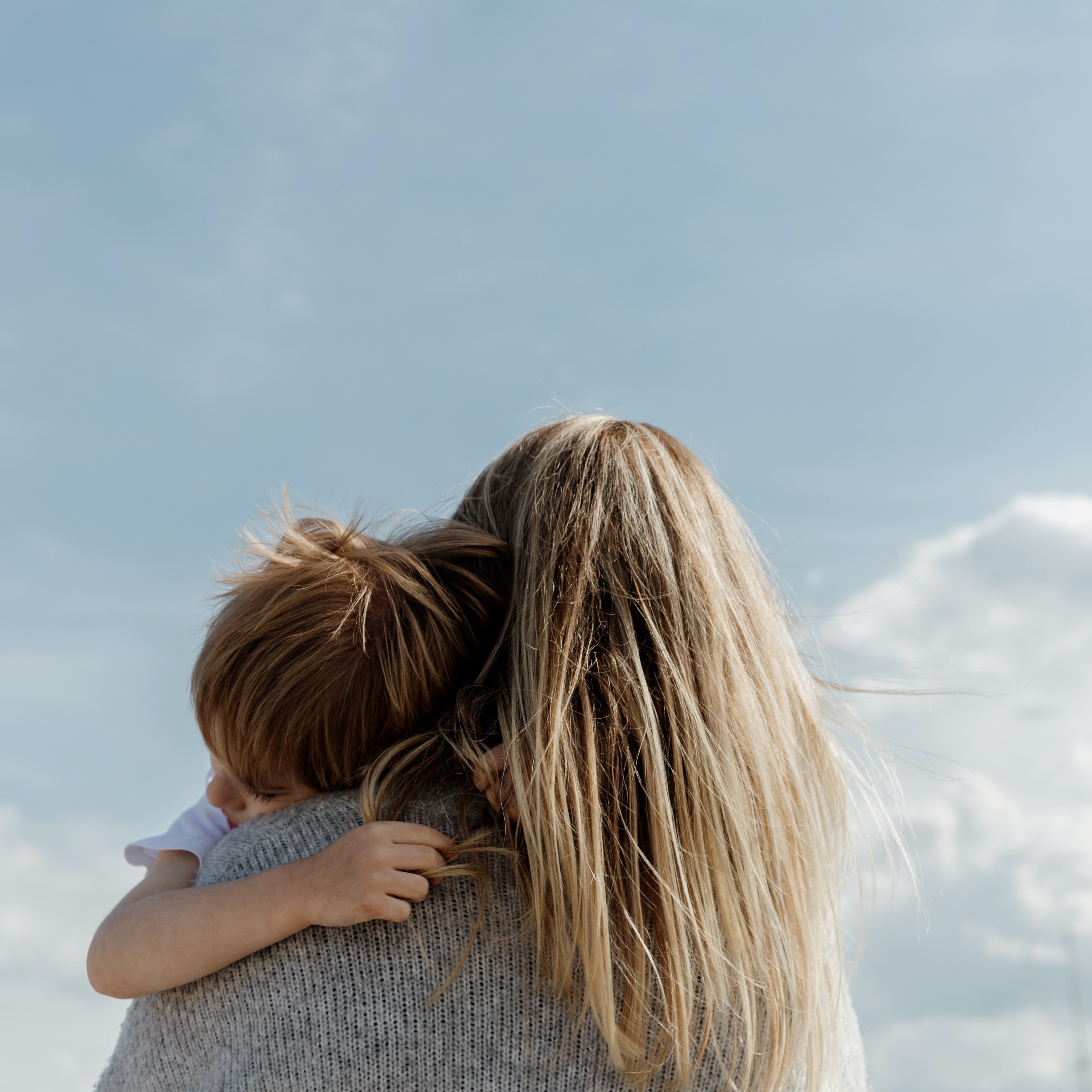 A person with long blonde hair holding a child. They are facing away from the camera and towards a blue sky.