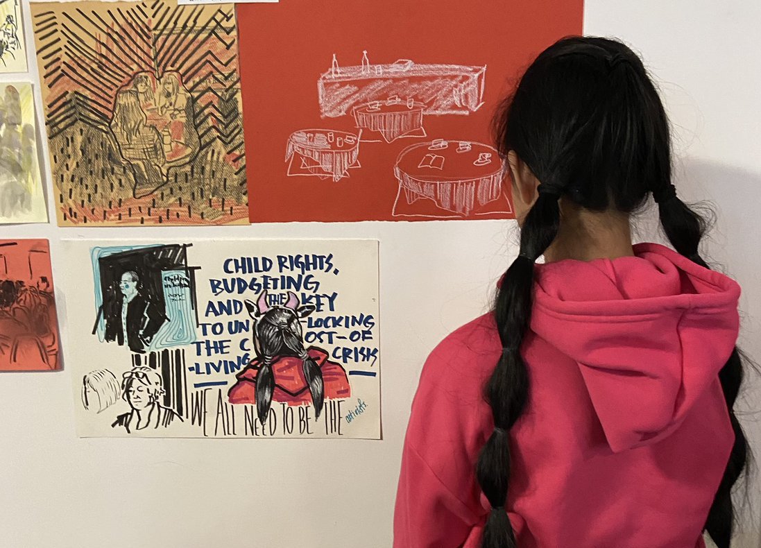 A girl with dark hair in two pony tails looks away from the camera and towards illustrations pinned to the wall. One of them shows the girl with ponytails, facing away from the illustrator.