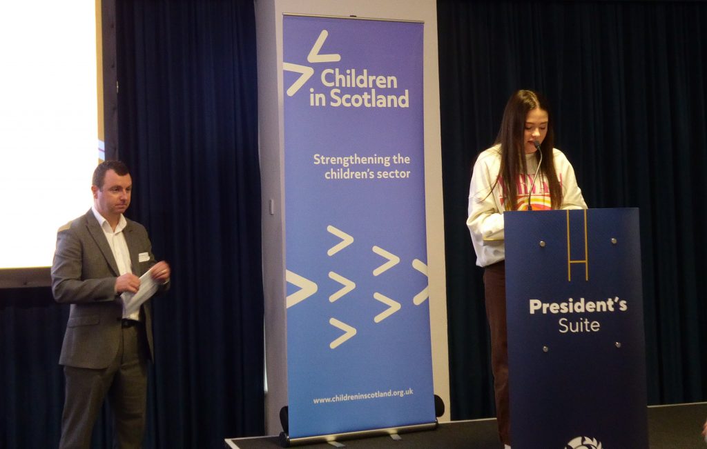 A girl standing behind a lectern, giving a speech. To her left is a man listening, and a sign that says Children in Scotland.