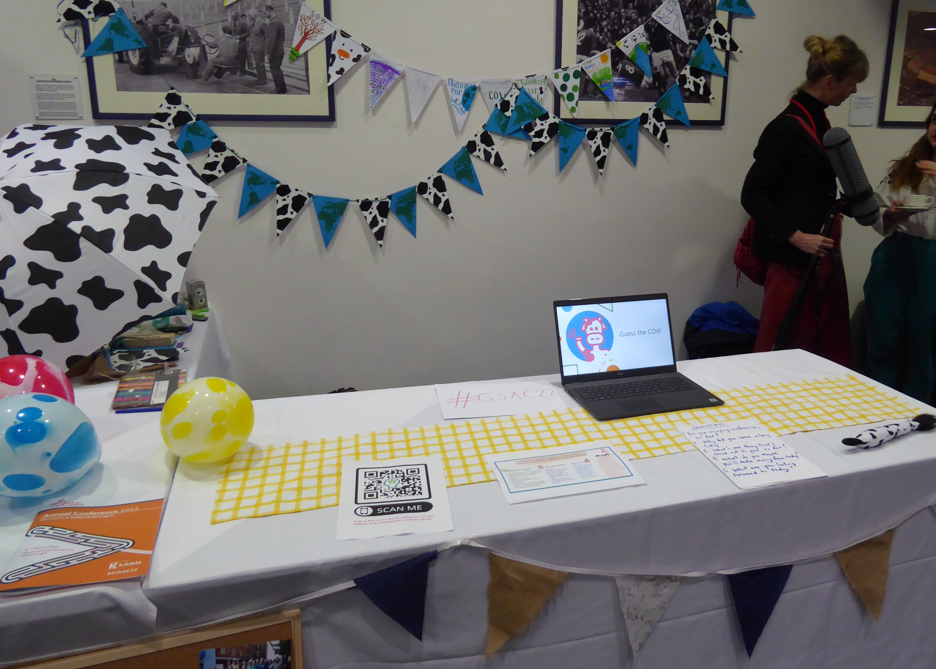 A table with a laptop open with 'Guess the cow' on it. On the table is an inflatable globe and a cow-print umbrella. There is cow-print bunting on the wall behind.