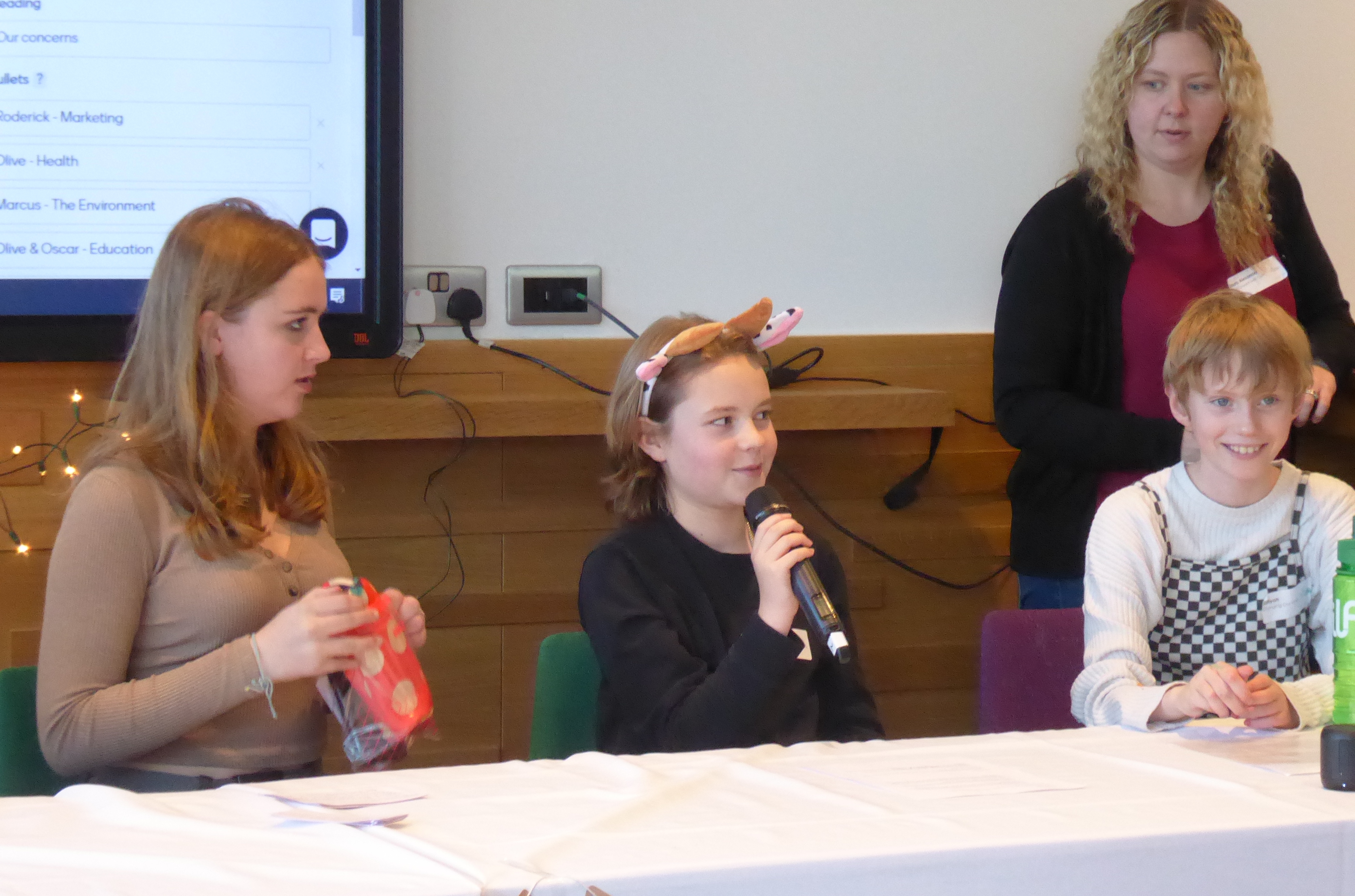 A girl wearing Cow ears holding a microphone, sitting behind a table with two other young people on either side. There is a woman standing behind, listening.