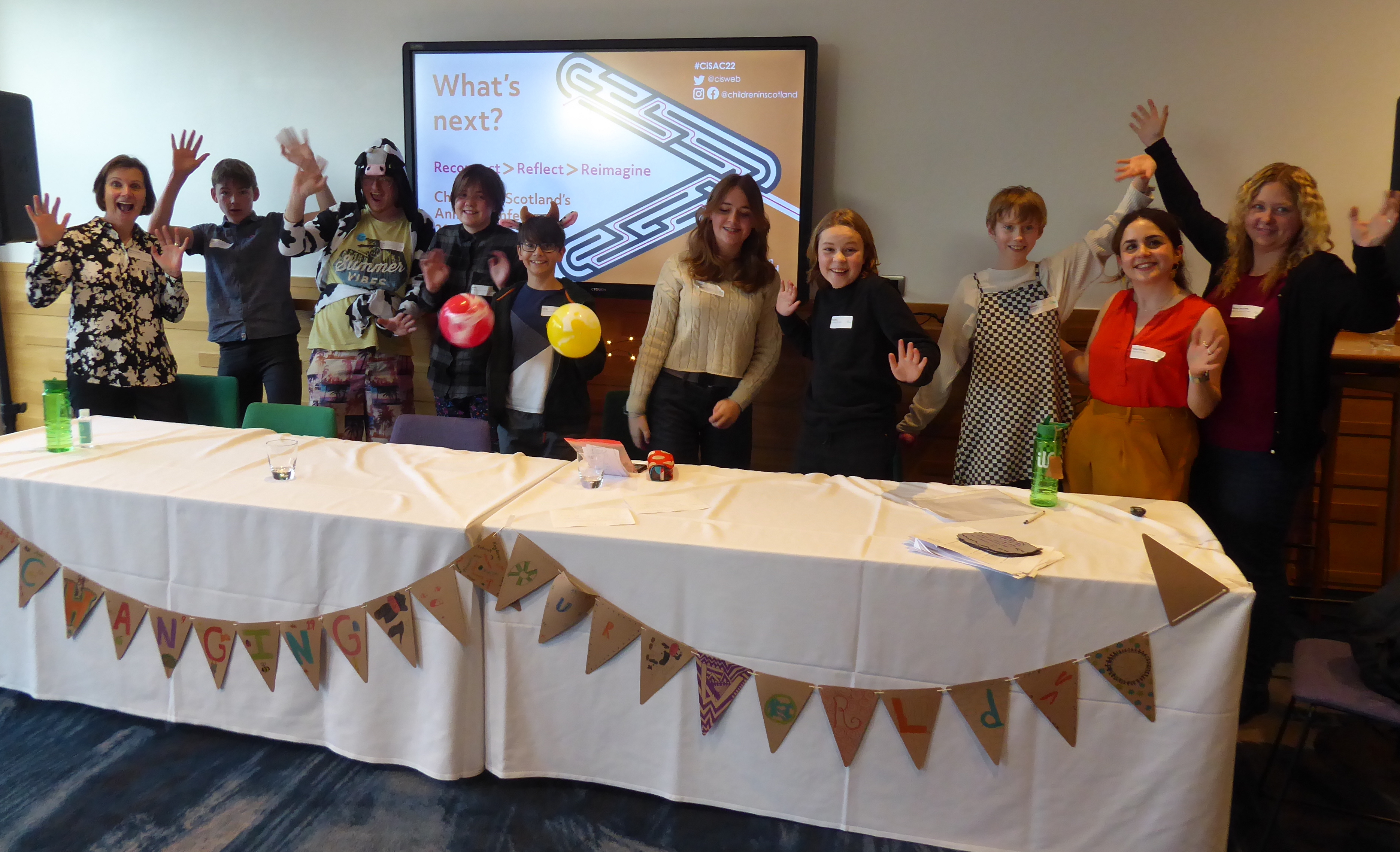 Adults and young people standing behind a table in front of a presentation screen. They have their arms up and are smiling.