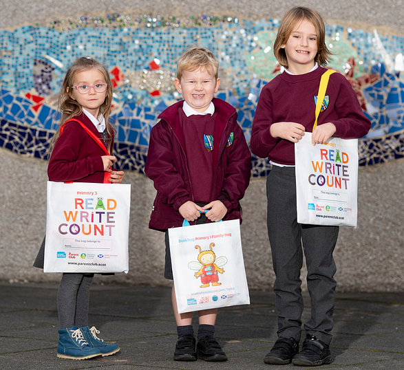Photo. Three primary school-aged children stand holding Read, Write, Count bags in front of a school mural.