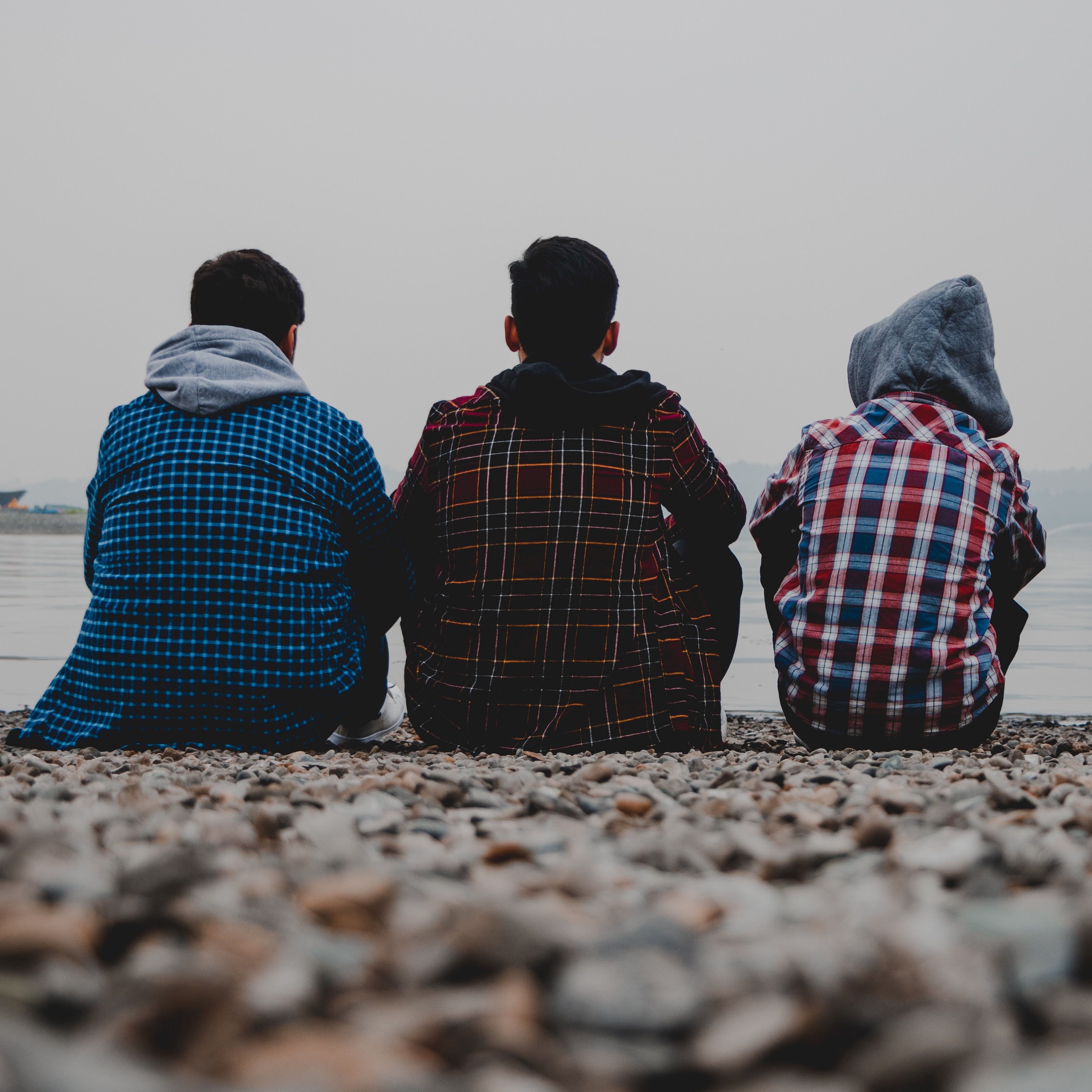 Three young people with short dark hair sitting on a rocky beach facing away from the camera. One has their hood up and all are wearing checked shirts.