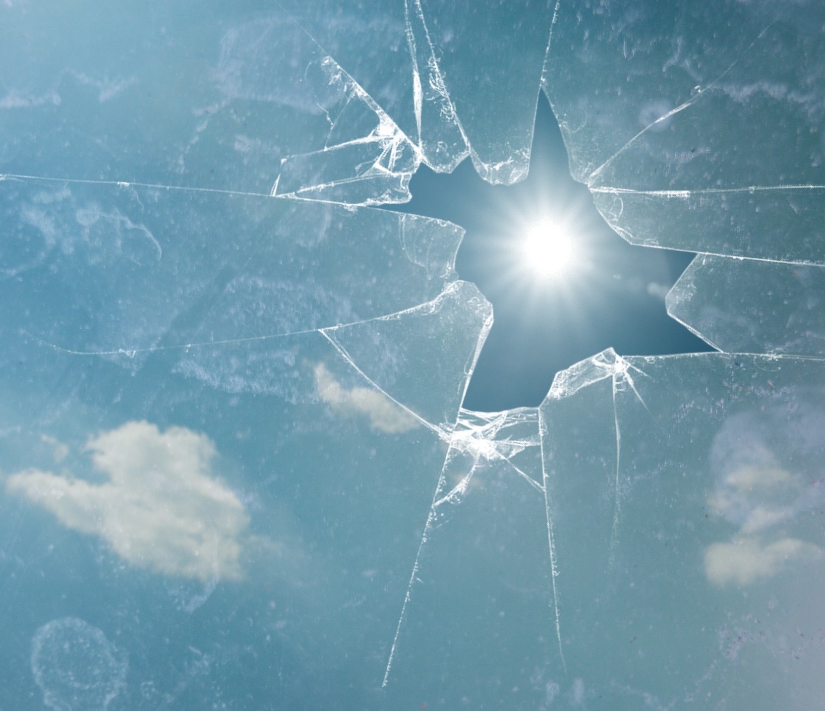 Photo. The sun shines brightly in a blue sky, viewed through a broken glass window.