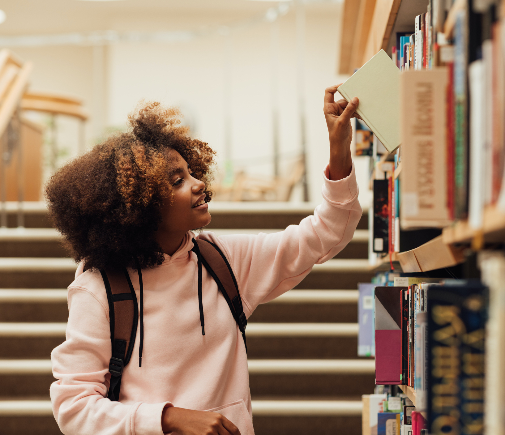 Photo. A girl is taking a book from a bookshelf in a library. She is wearing a pink hooded top and wearing a rucksack.